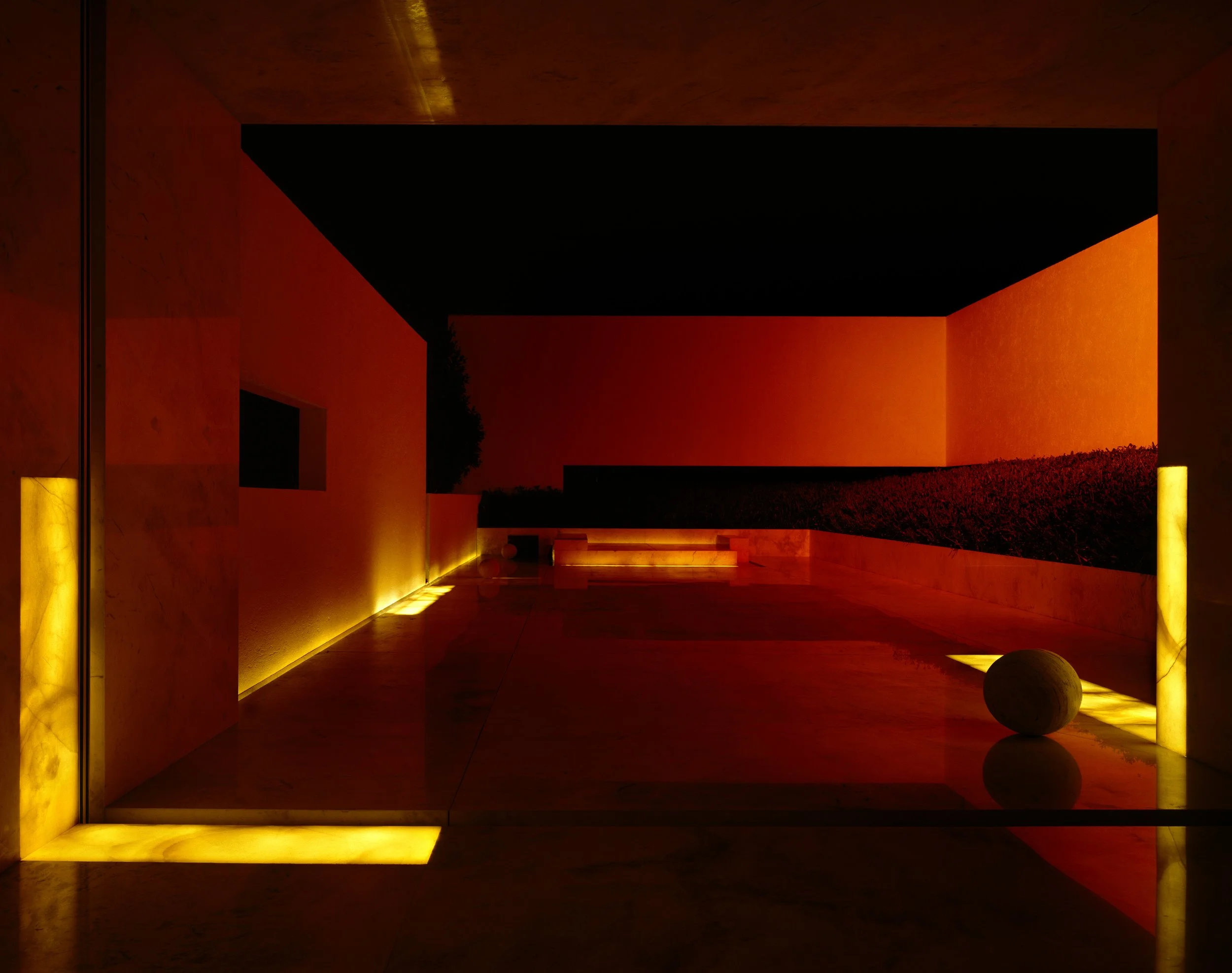 Vista nocturna de un edificio moderno con iluminación cálida, paredes de color terracota y columnas amarillas, con reflejo en el suelo de piedra pulida.