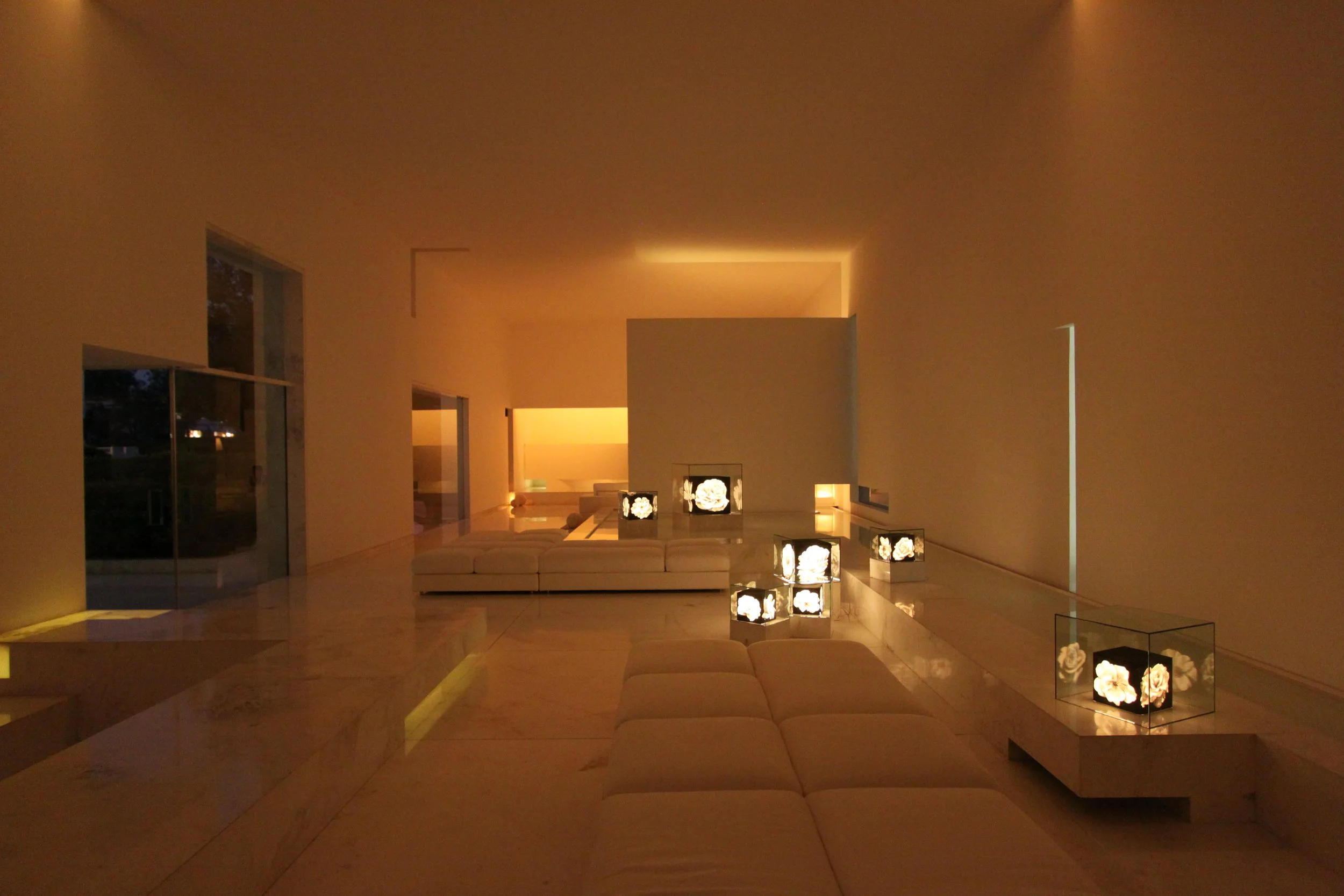 Interior de una sala moderna con iluminación cálida, sillones blancos y lámparas decorativas con flores.