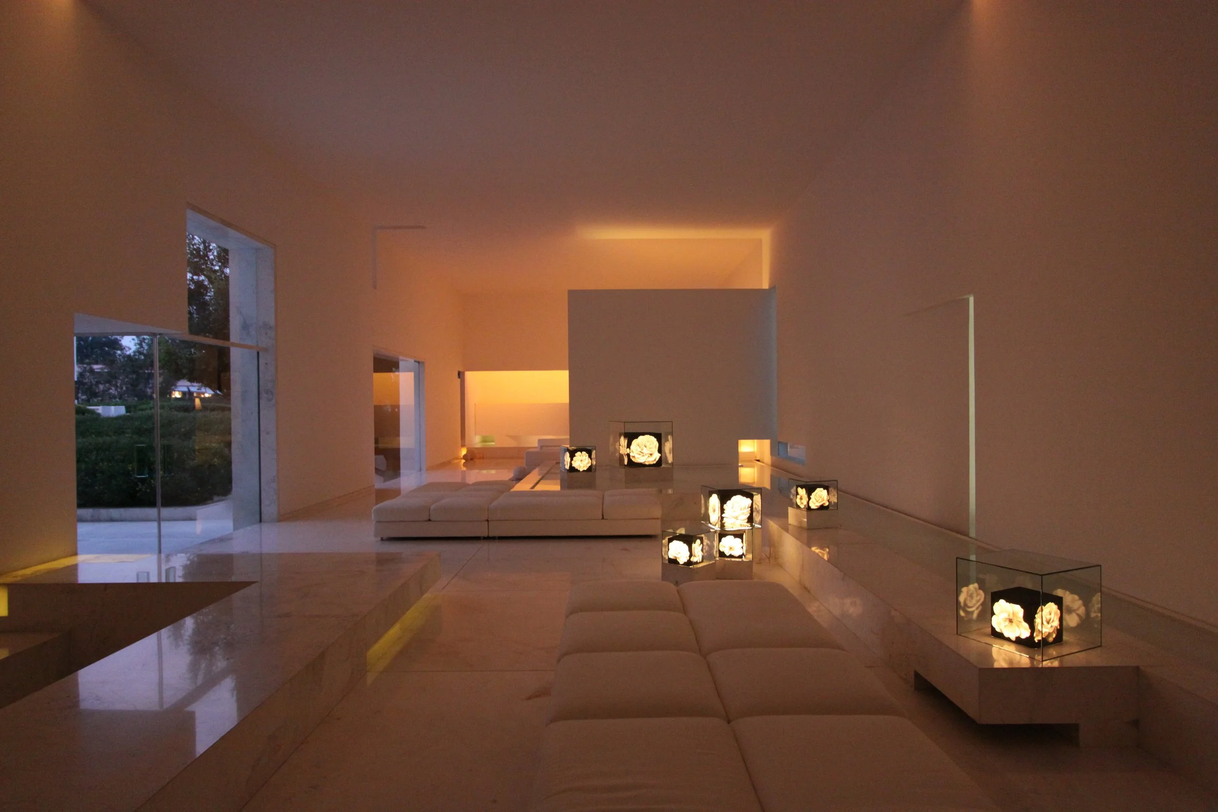 Interior de una sala moderna con iluminación cálida, sillones beige, mesas con decoraciones de flores en caja de luces