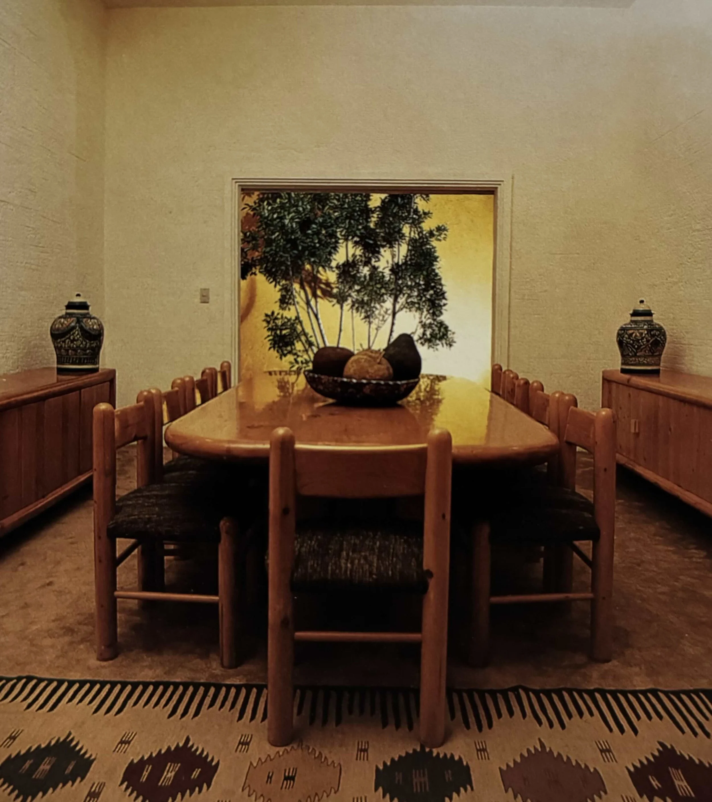 Sala de comedor con mesa de madera y sillas, decorada con jarrones con motivos en ambos lados y un cuadro con árbol en el fondo.