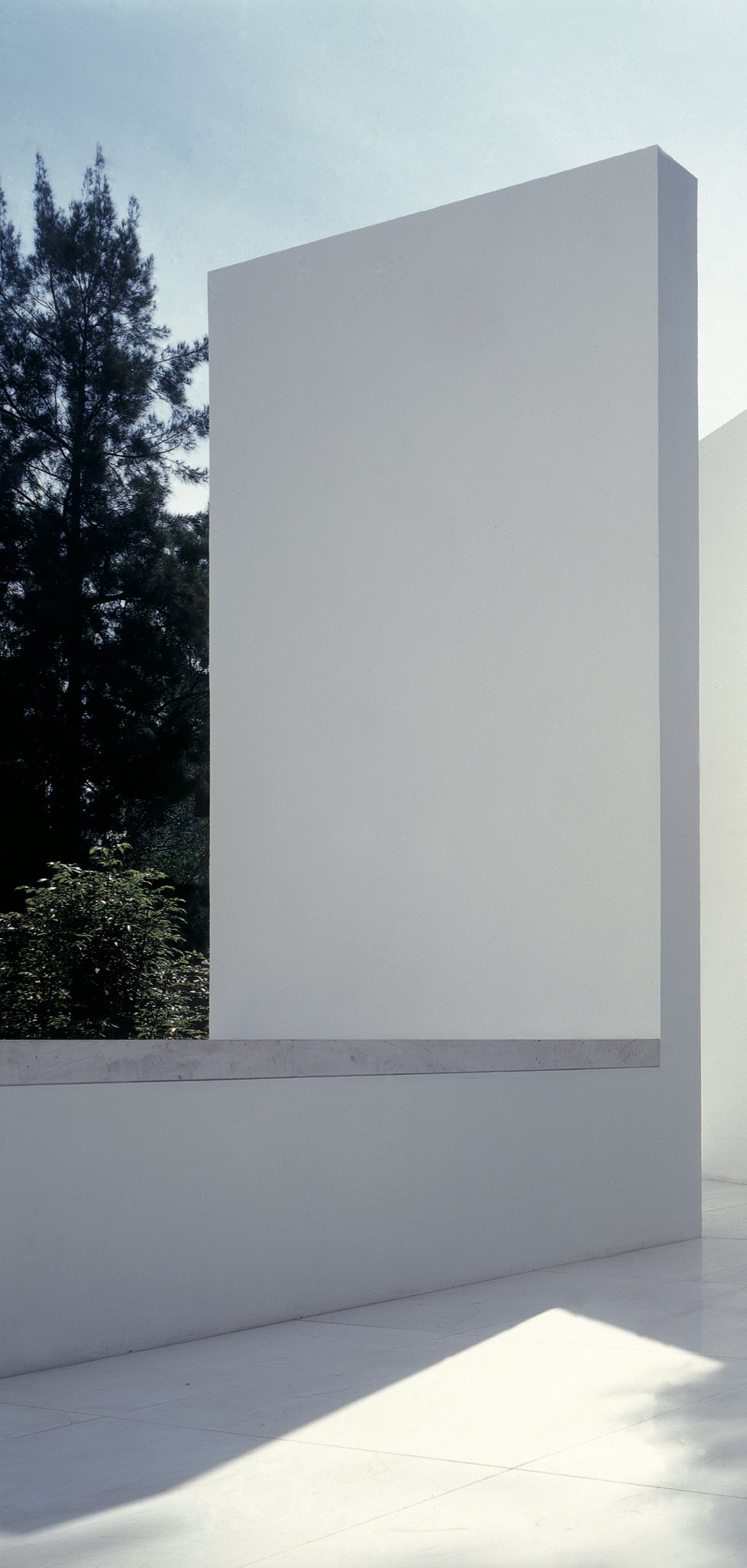 fachada de un edificio blanco minimalista con sombra en el suelo y árboles verdes al fondo