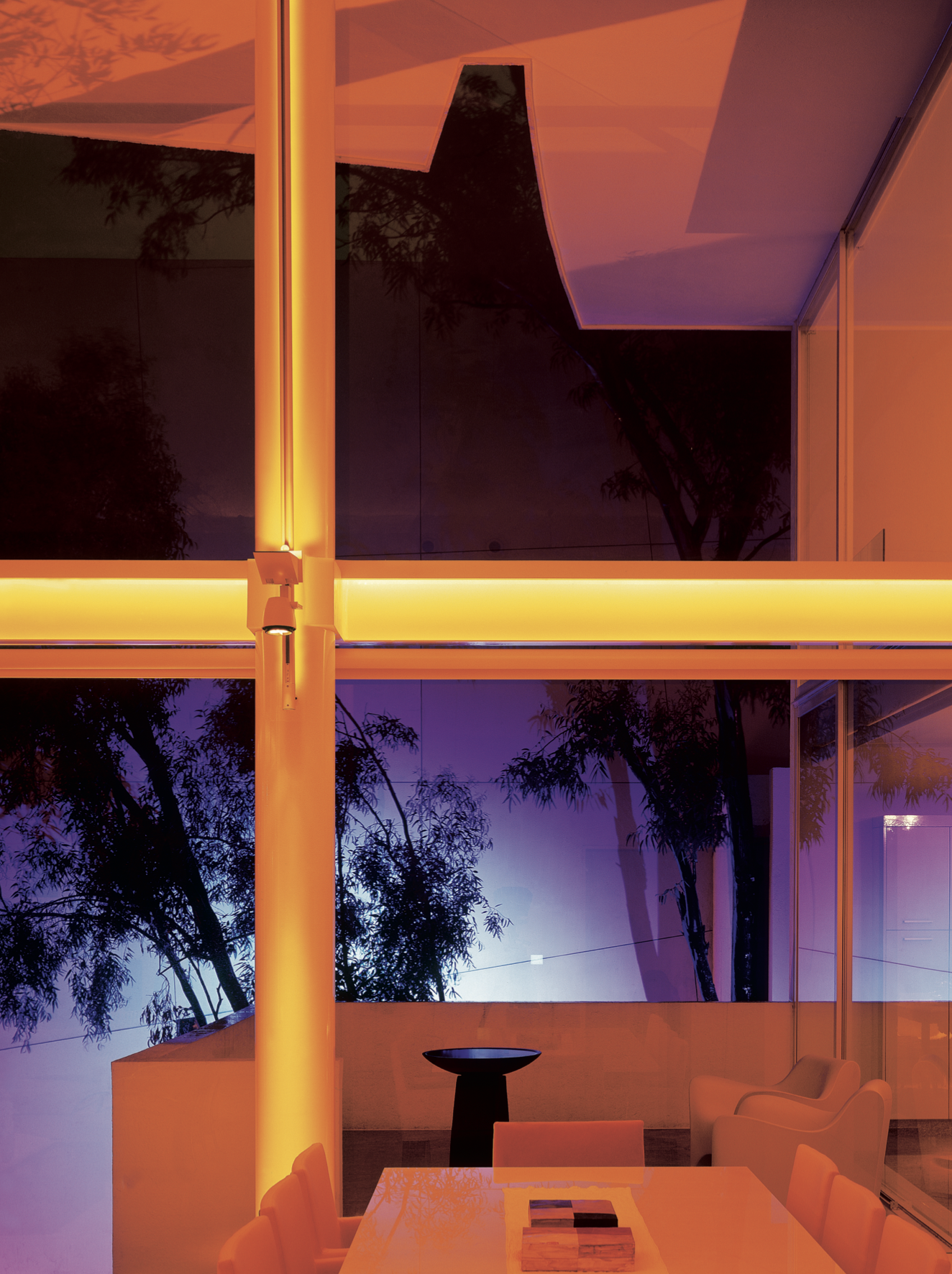 Vista interior de un espacio con ventanas grandes y árboles visibles afuera, iluminado con luz naranja y azul, con mobiliario moderno incluyendo una mesa y sillas.