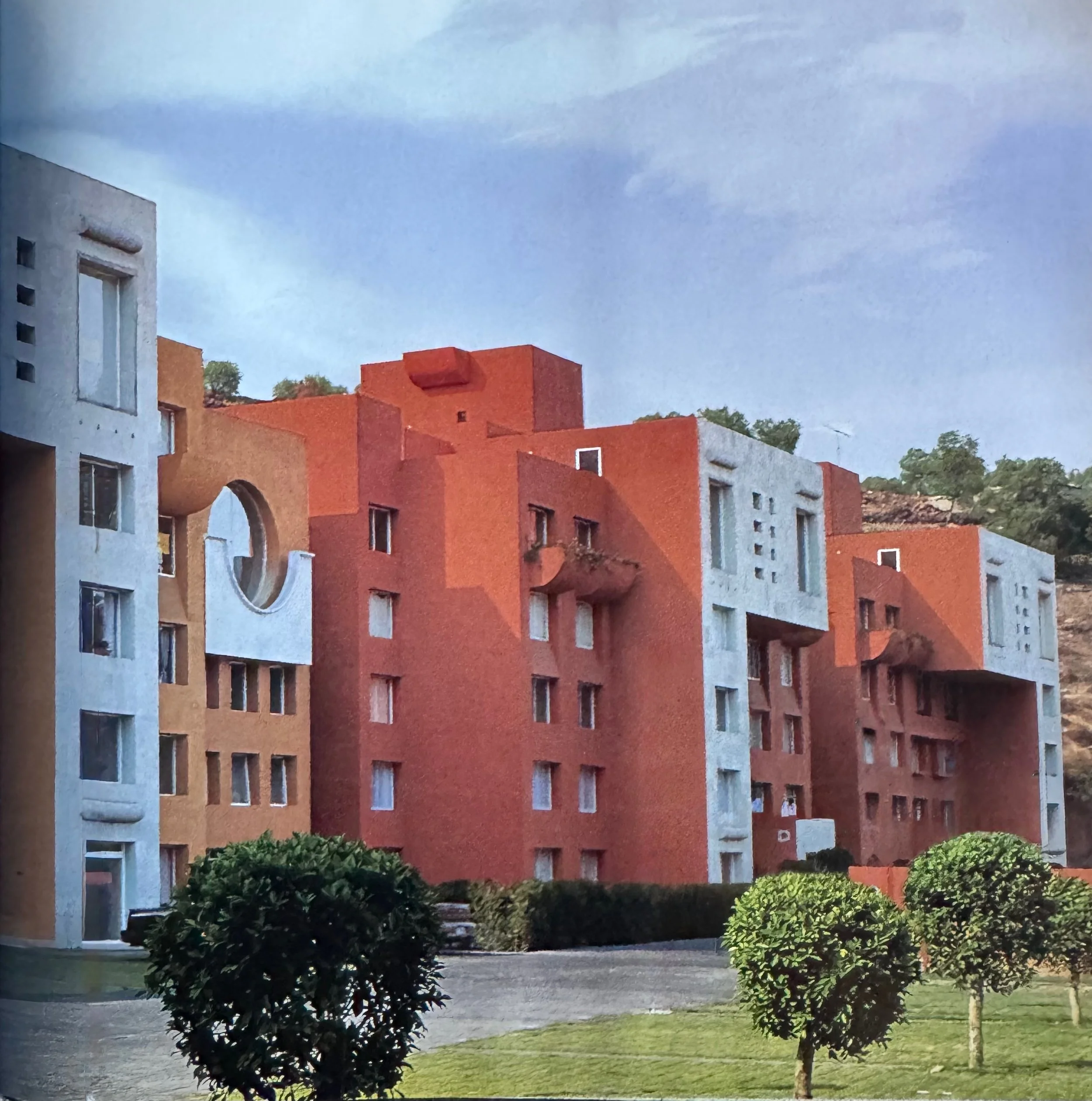 Edificio de apartamentos de color gris y rojo con árboles y arbustos en primer plano, cielo nublado.