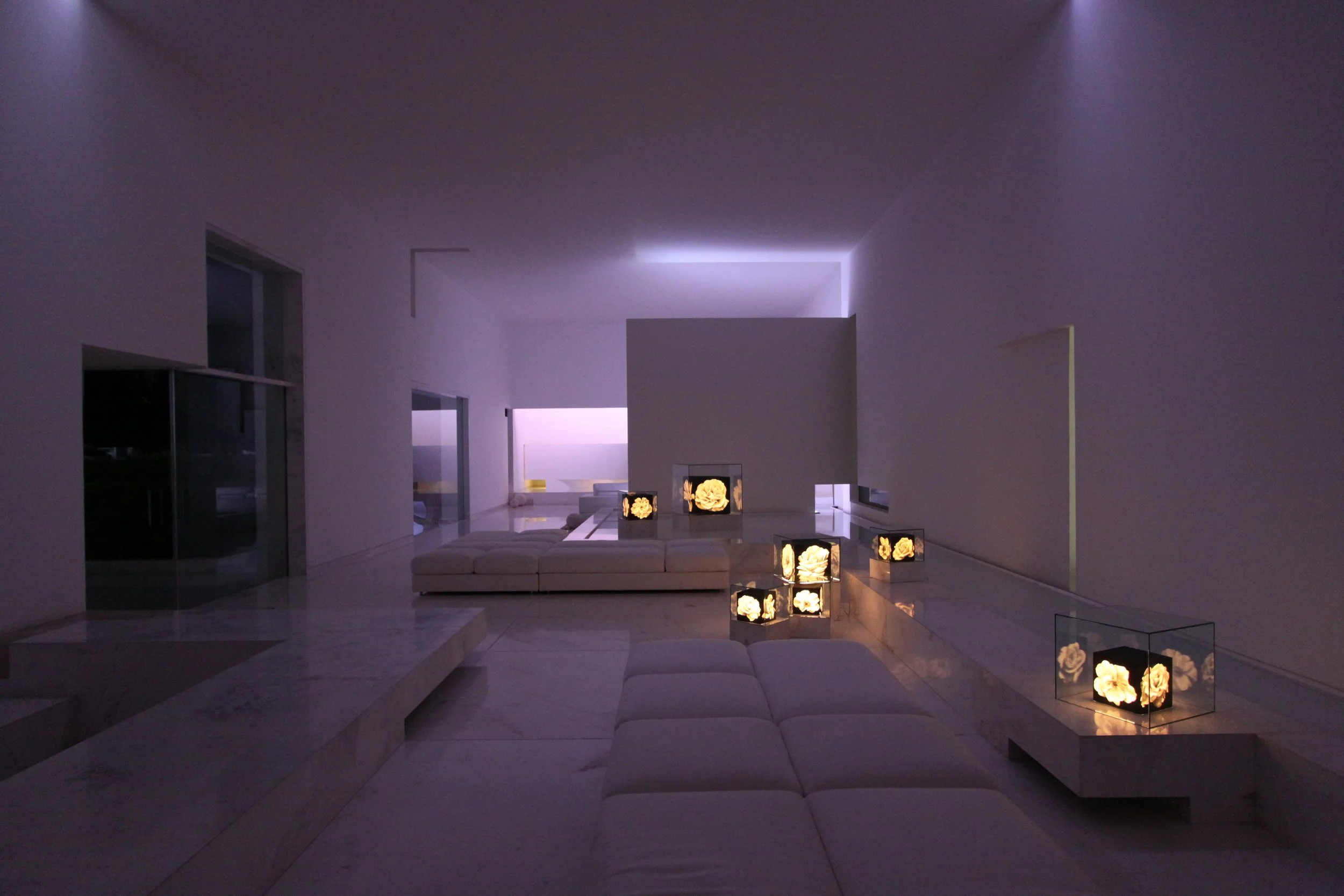 Interior moderno con iluminación tenue, sofá blanco, mesas con lámparas con flores en su interior, paredes blancas y ventanas grandes. Architecture & Design by Miguel Ángel Aragonés | Taller Aragonés. Photography by Joe Fletcher