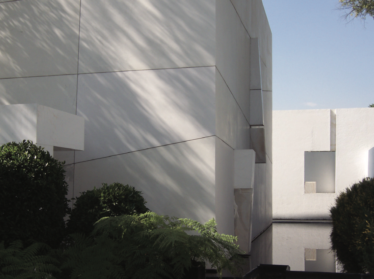 Edificio moderno de paredes blancas con ventanas cuadradas, acompañado de arbustos y plantas verdes al frente y sombra de árboles sobre la pared.