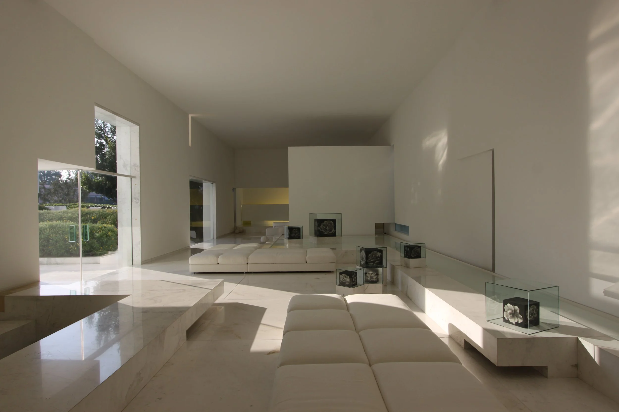 Interior de una sala moderna con paredes blancas y ventanas grandes que permiten la entrada de luz natural, pisos de mármol y muebles blancos, decorada con obras de arte en exhibición.