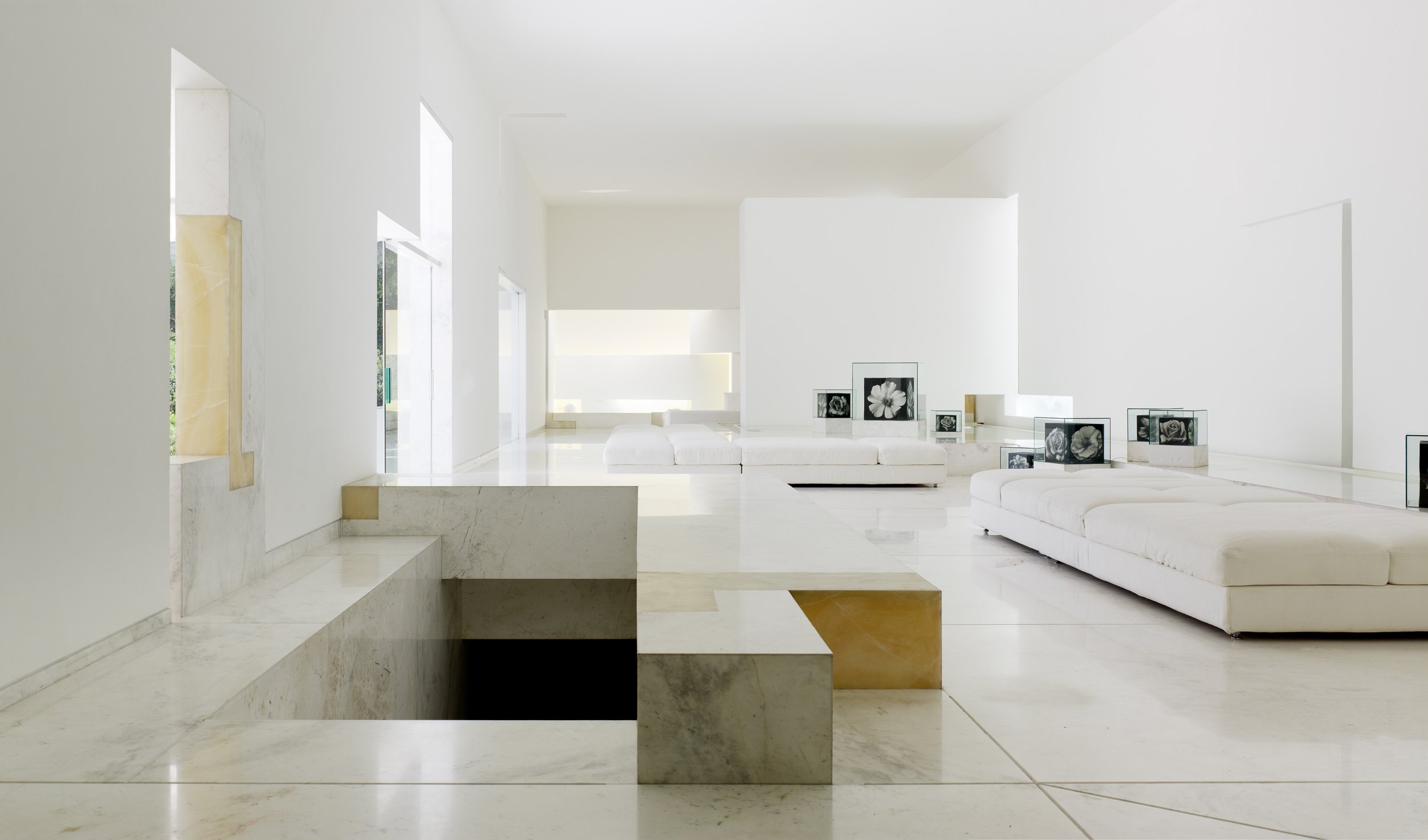 Interior de una sala minimalista con paredes blancas, sillones blancos y varias fotografías en cajas de vidrio con flores en exhibición. Architecture & Design by Miguel Ángel Aragonés | Taller Aragonés. Photography by Joe Fletcher