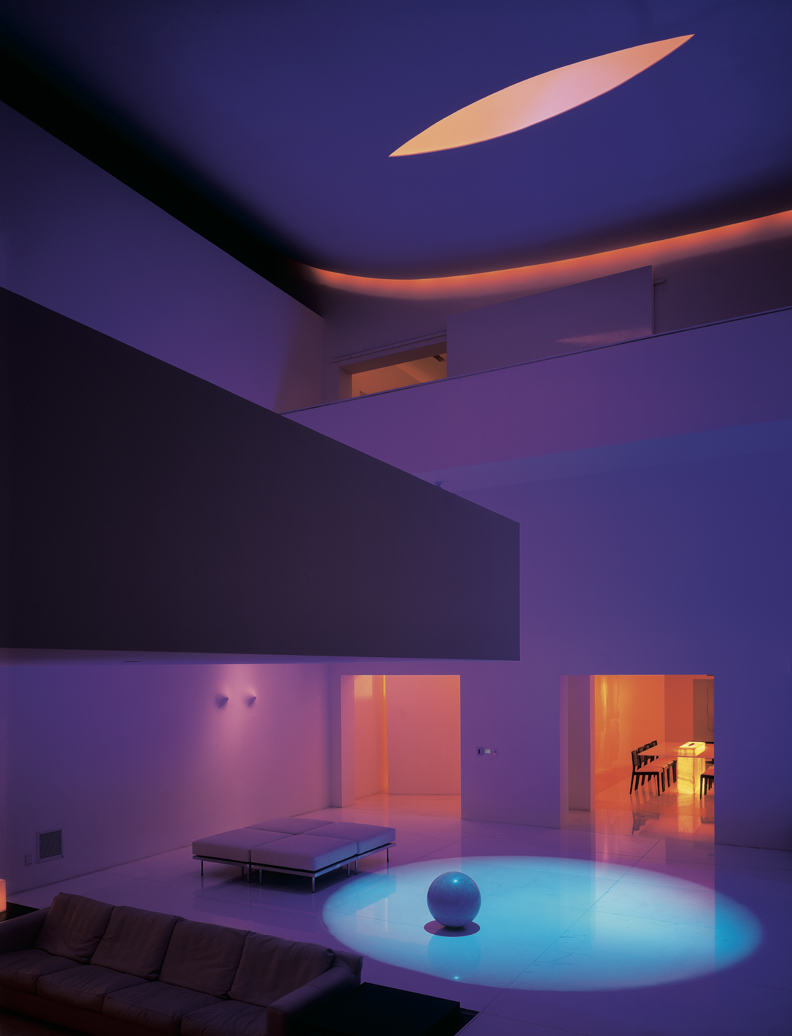 Interior moderno con iluminación de colores en tonos morados, azules y naranjas, incluye sofá gris, mesa de centro, pelota grande decorativa, y comedor con sillas en una sala amplia y luminosa. Chromatic architecture in Eucaliptos 65 by Miguel Ángel 