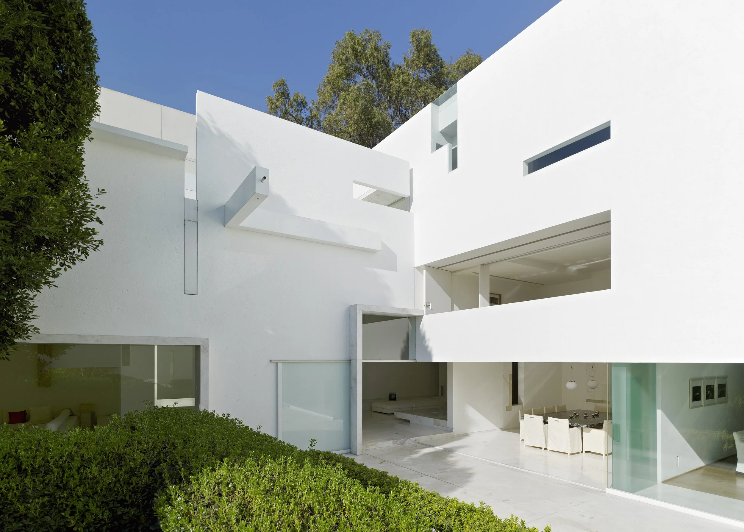 Vista de una casa moderna de paredes blancas, con espacio de terraza y áreas abiertas, rodeada de vegetación y árboles, con cielo claro. Architecture & Design by Miguel Ángel Aragonés | Taller Aragonés. Photography by Joe Fletcher
