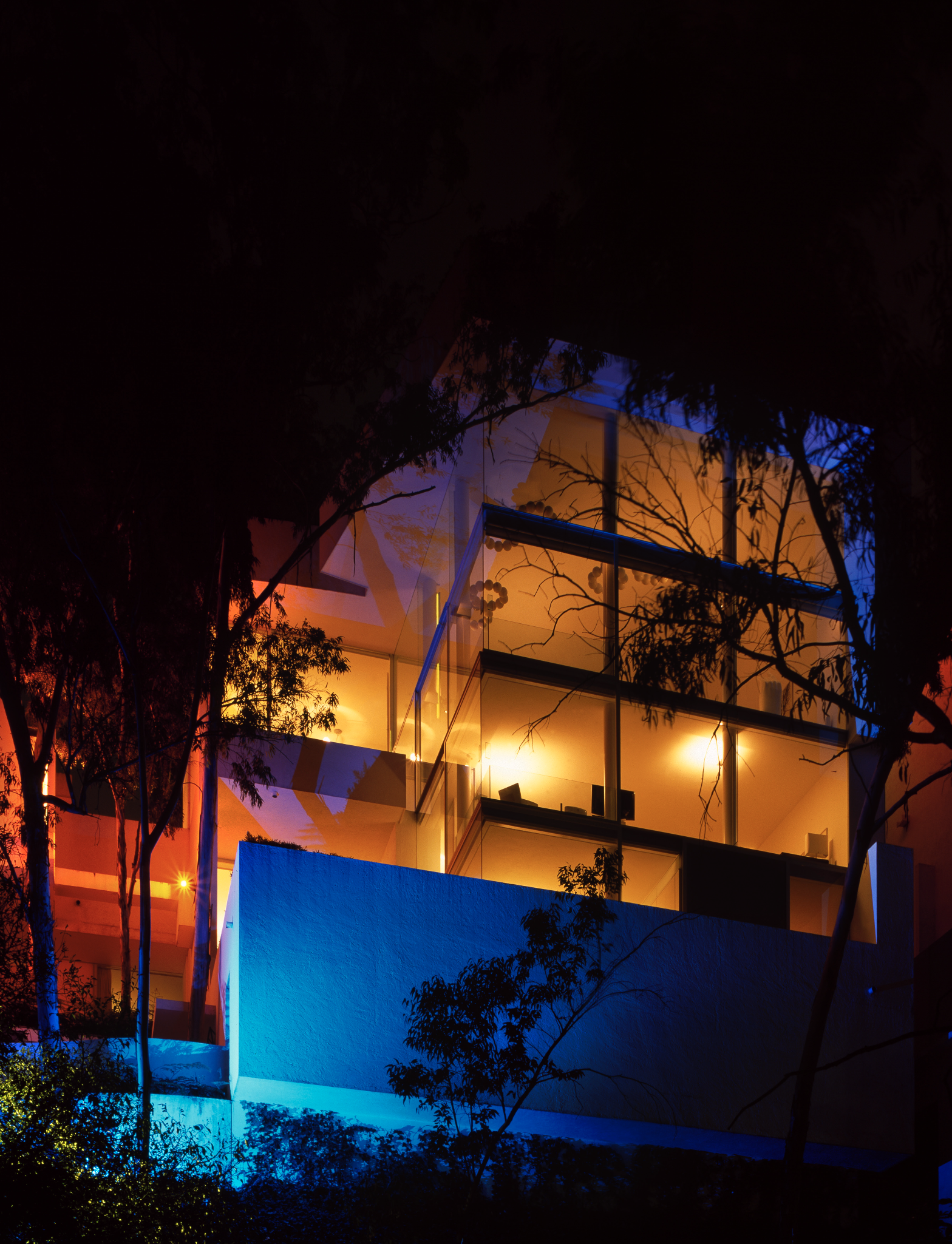 Edificio moderno con fachadas de vidrios reflectantes iluminadas con luces de colores azul y naranja, rodeado de árboles y vegetación en la noche.