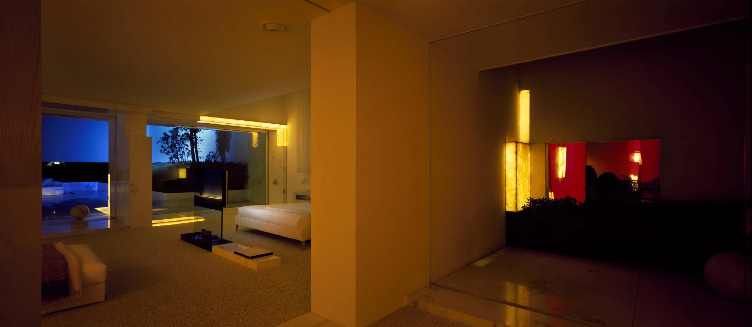 Interior de una habitación moderna con iluminación cálida, vista a un área exterior con piscina y árboles, con ventanas grandes y muebles blancos.