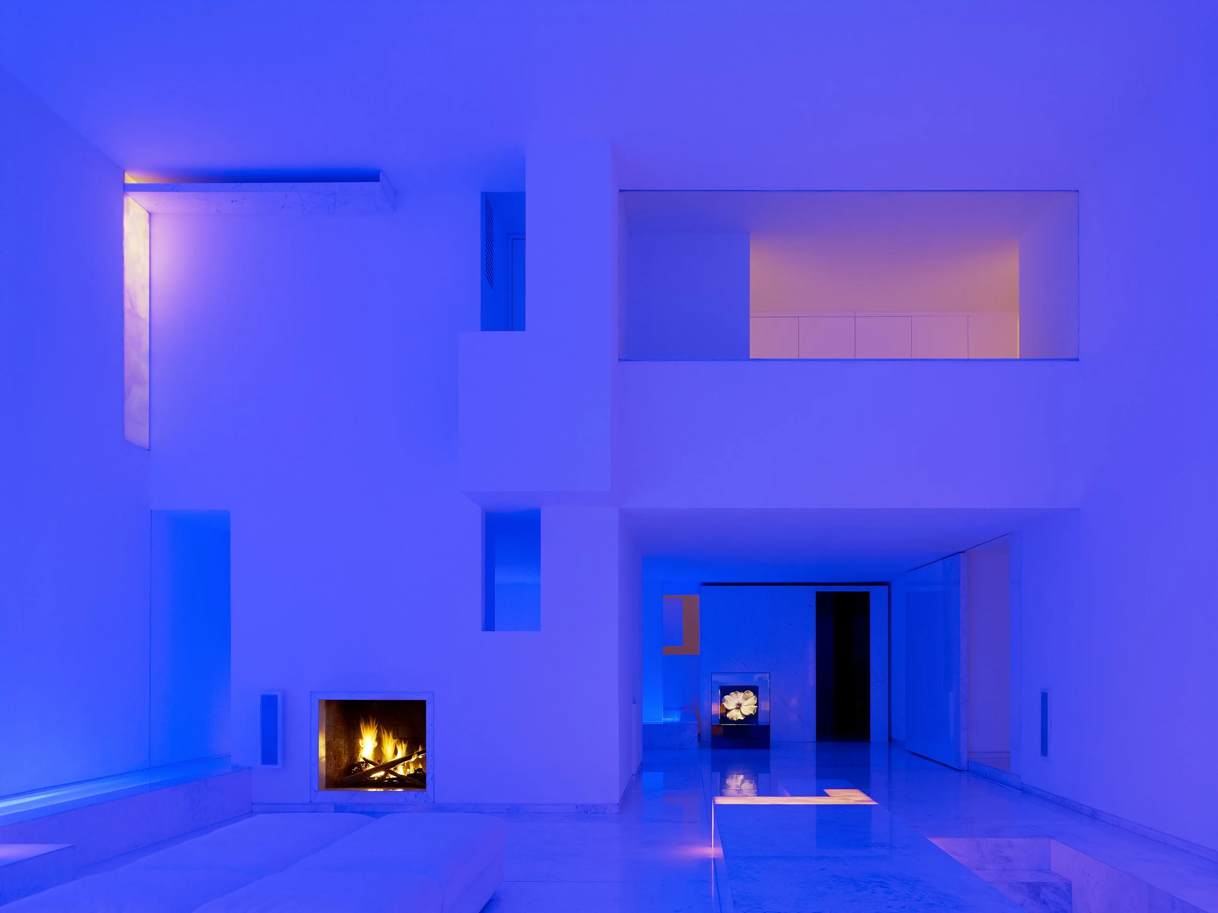 Sala moderna con iluminación azul, chimenea encendida, en la pared hay varias aberturas y una pantalla con flores. Architecture & Design by Miguel Ángel Aragonés | Taller Aragonés. Photography by Joe Fletcher