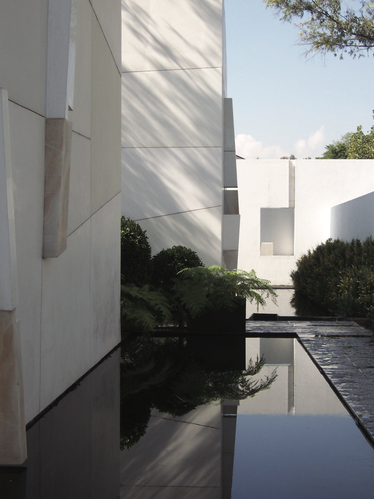 Construcción moderna de paredes blancas y ventanas cuadradas, con un reflejo en un estanque de agua, y vegetación en el entorno, bajo un cielo despejado.