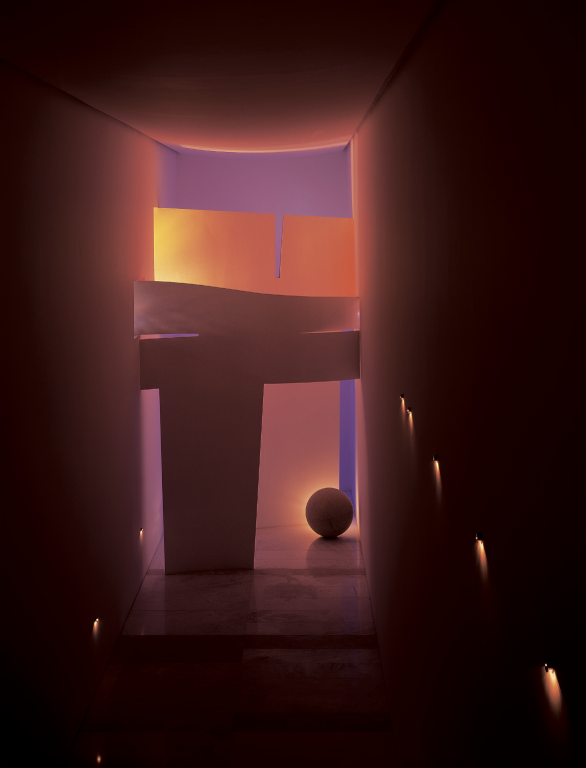 Pasillo oscuro con luces a lo largo de las paredes, en el fondo hay una escultura blanca y una esfera, con iluminación en tonos cálidos y lilas. Eucaliptos 65, Architecture & Design by Rafael Aragonés Caballero & Miguel Ángel Aragonés | Taller Aragon