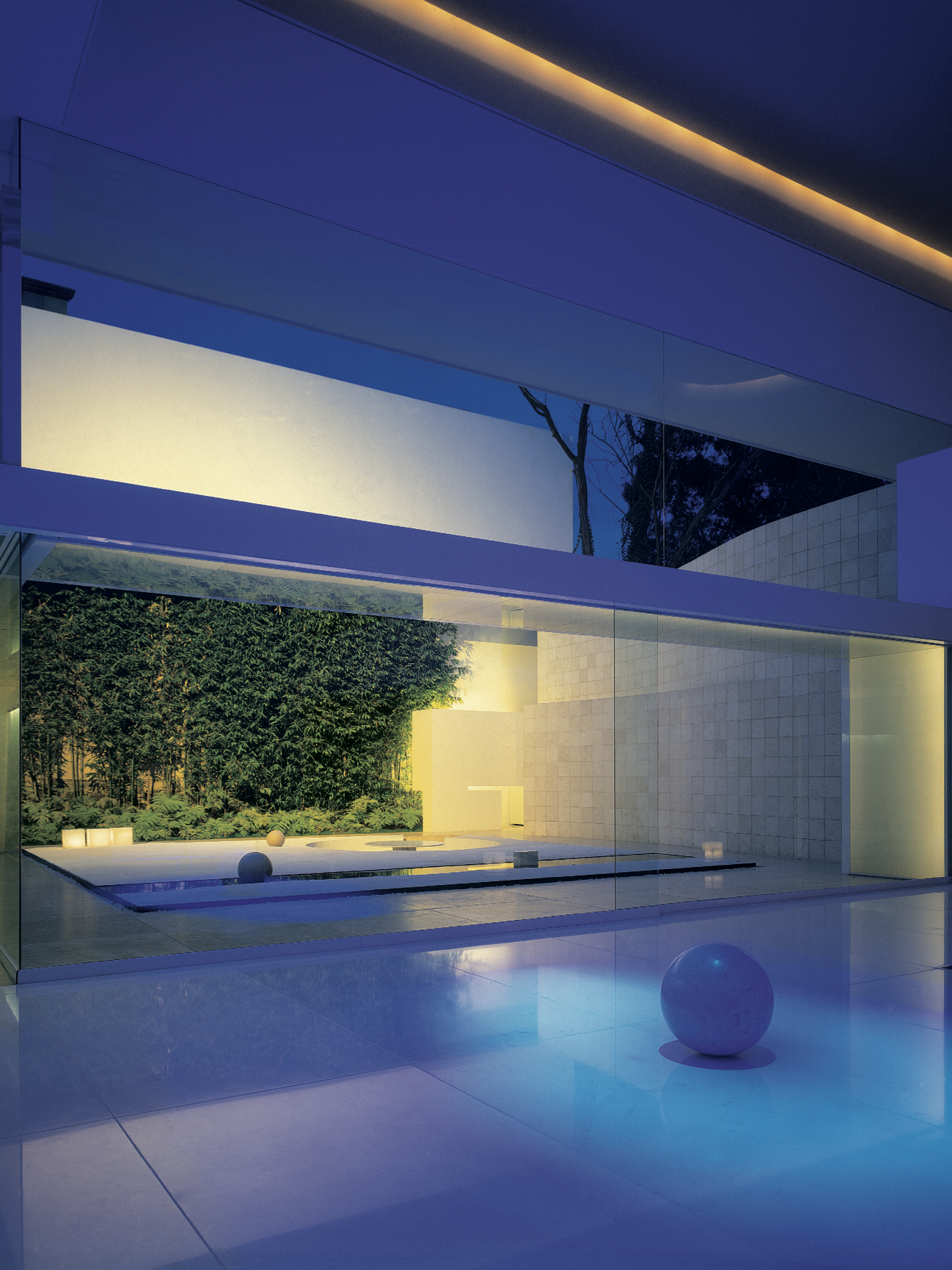 Exterior de una casa moderna con paredes de vidrio y un jardín con césped, árboles y bolas decorativas, iluminada con luces cálidas y de colores, en la noche. Eucaliptos 65, Architecture & Design by Rafael Aragonés Caballero & Miguel Ángel Aragonés |