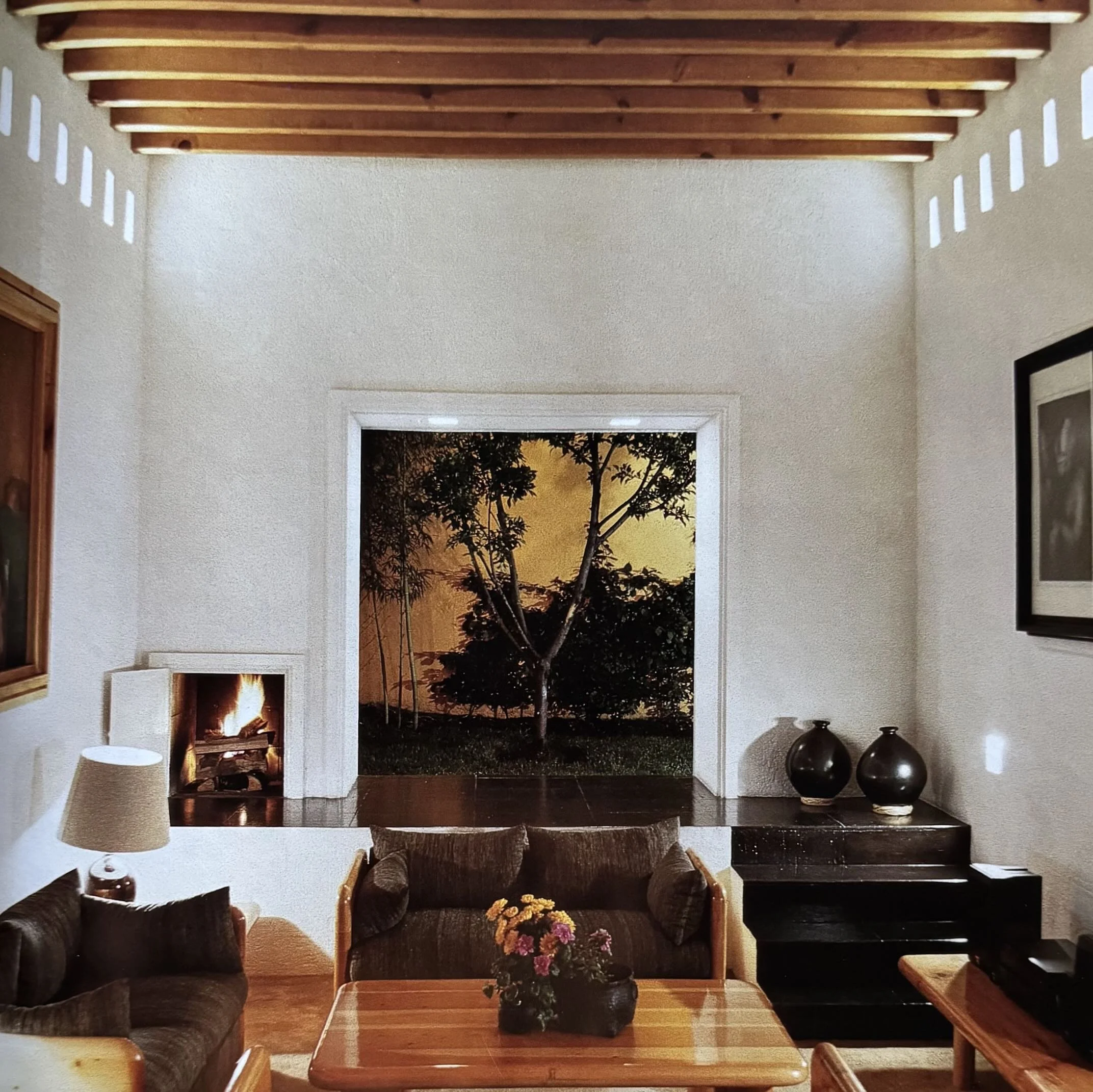 Sala de estar con sillones, mesas de madera, una chimenea, jarrones grandes y una vista de un atardecer con árboles y cielo nublado.