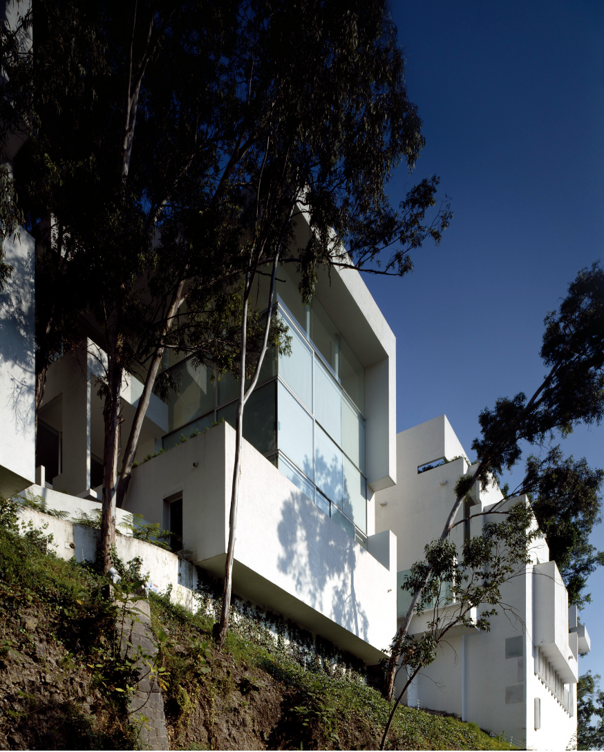 Casa moderna blanca con grandes ventanas de cristal, ubicada en una colina con árboles y vegetación alrededor, bajo un cielo despejado y azul.