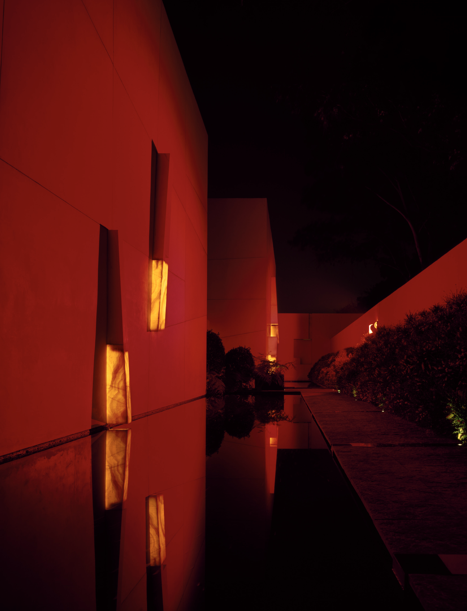 Una vista nocturna de un edificio con paredes rojas, iluminadas por luces amarillas, y un camino rodeado de arbustos y árboles oscuros.