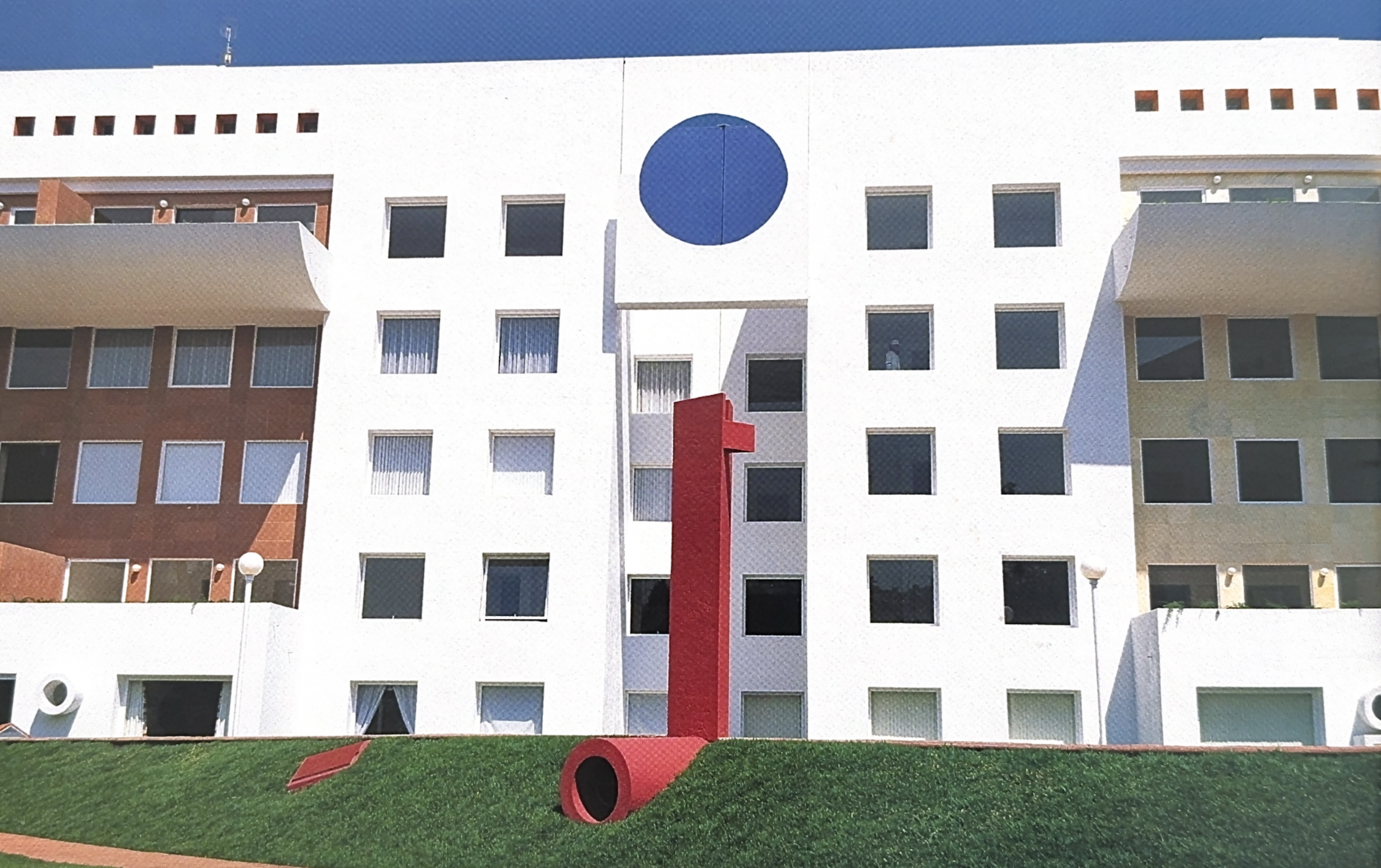 Edificio de apartamentos de varios pisos con ventanas rectangulares y una esfera azul en la fachada. En primer plano, una escultura roja en forma de tubo curvado. La escena está en un área con césped bien cuidado.