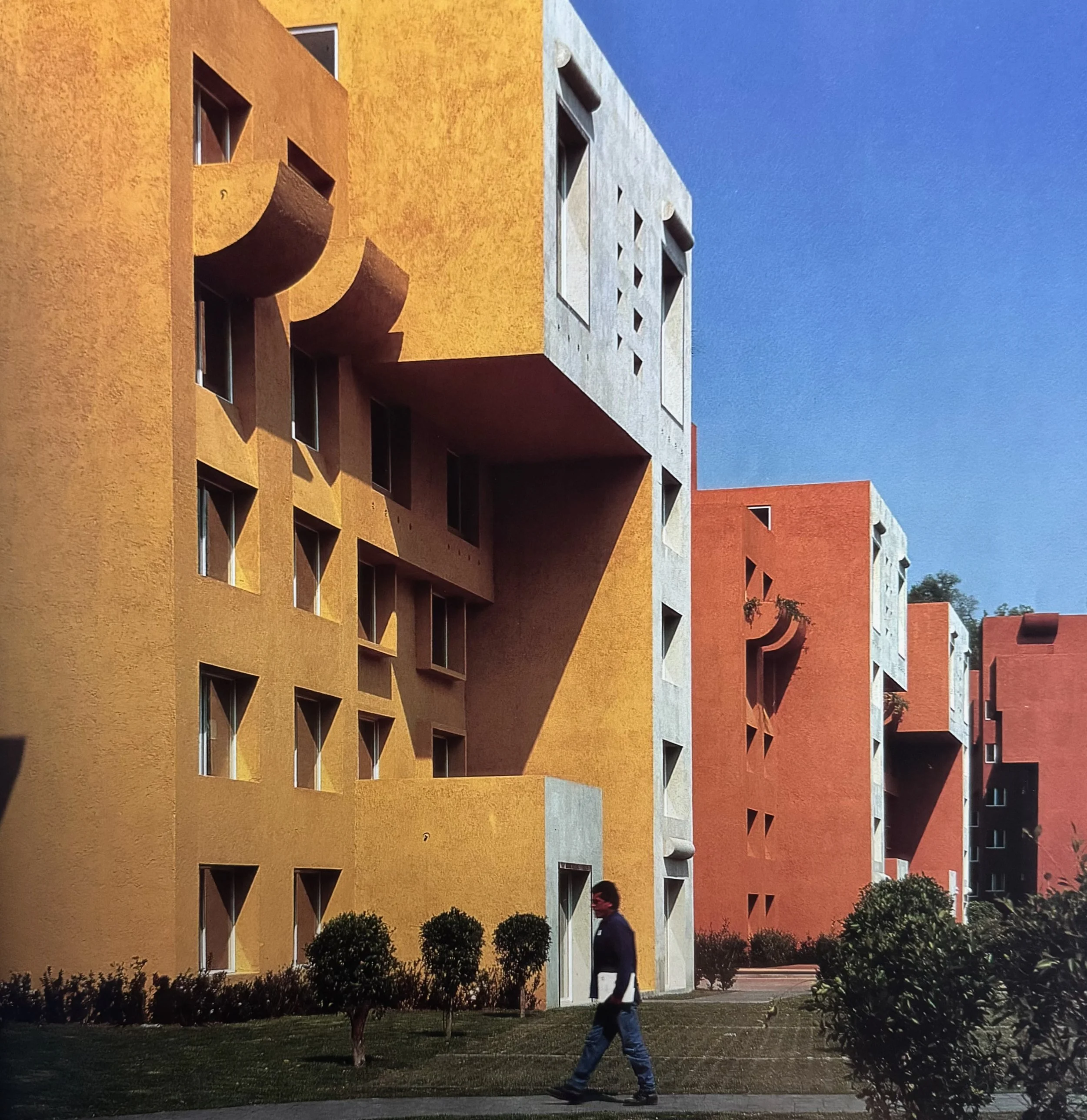 Edificio de apartamentos con paredes en colores naranja, gris y blanco, con ventanas y áreas de reflexión, rodeado de árboles y arbustos en la noche con cielo claro.