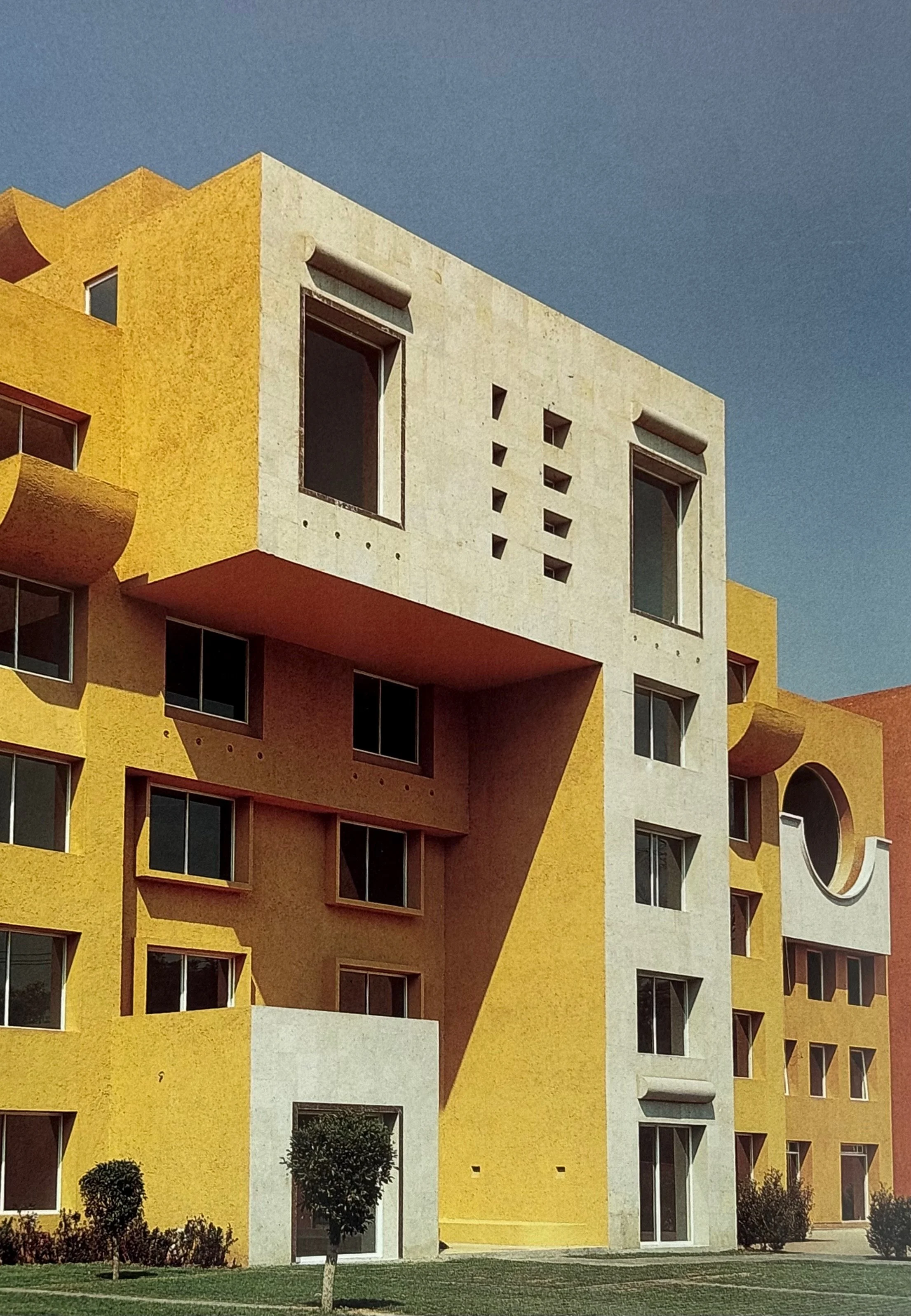 Edificio moderno de colores amarillo y blanco con ventanas grandes y formas arquitectónicas distintivas, en un entorno con césped y pequeños árboles.