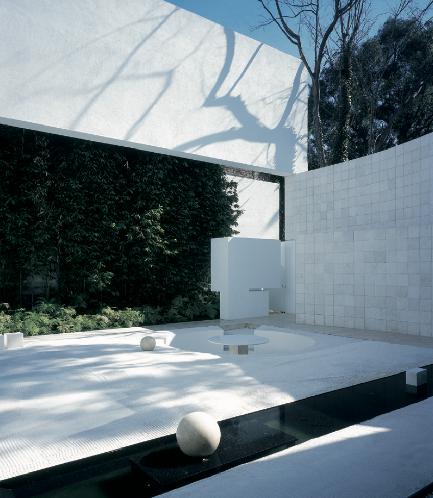 Un patio minimalista con paredes blancas, vegetación y sombras de árboles. Incluye bolas decorativas y una estructura moderna de forma rectangular y curva. Eucaliptos 65, Architecture & Design by Rafael Aragonés Caballero & Miguel Ángel Aragonés | Ta