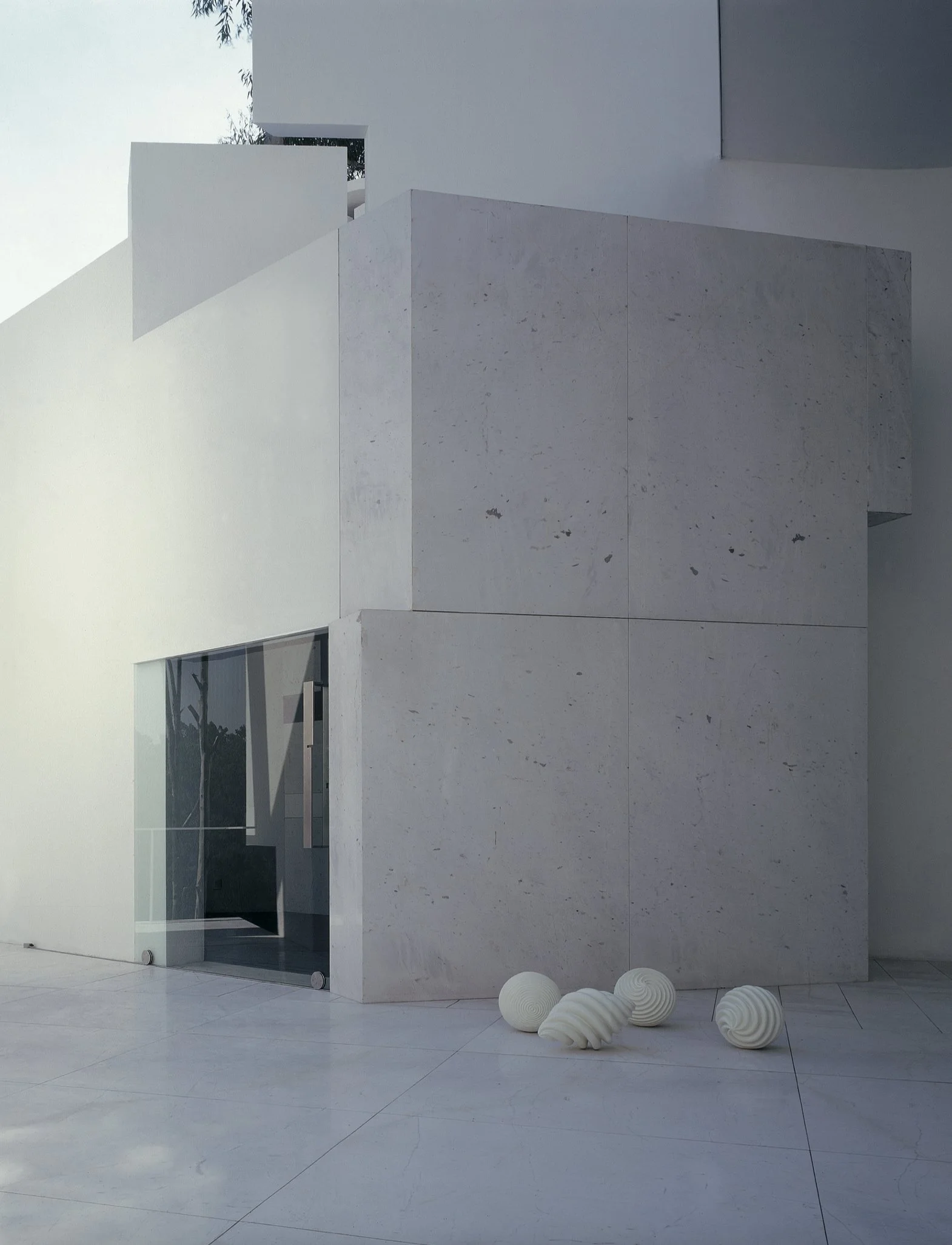 Vista exterior de un edificio blanco con paredes modernas y ventanas rectangulares, decorado con esculturas blancas en forma de conchas en el suelo.