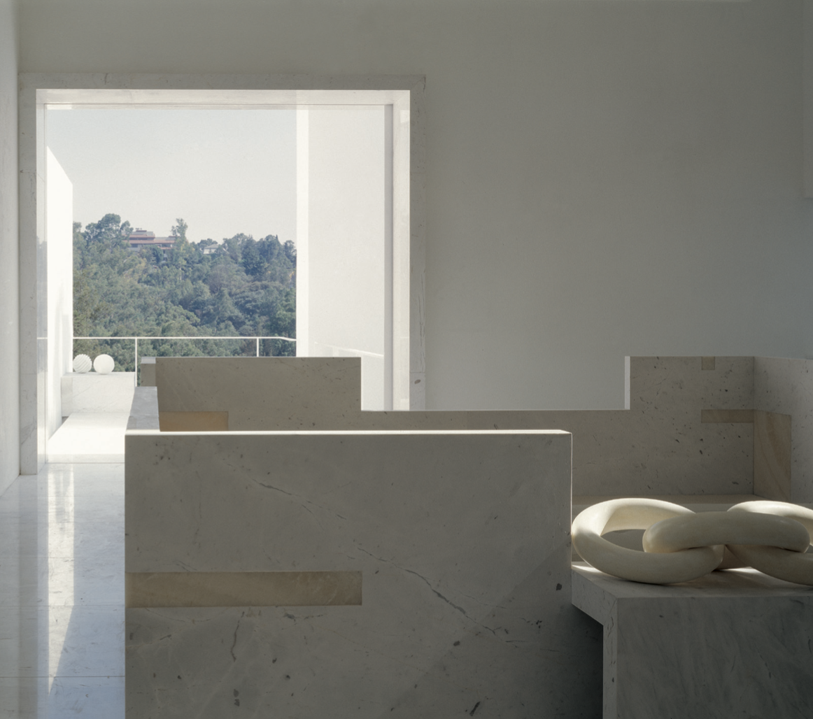 Interior de un espacio minimalista con paredes y muebles de mármol, con una ventana grande que muestra una vista de vegetación y colinas.
