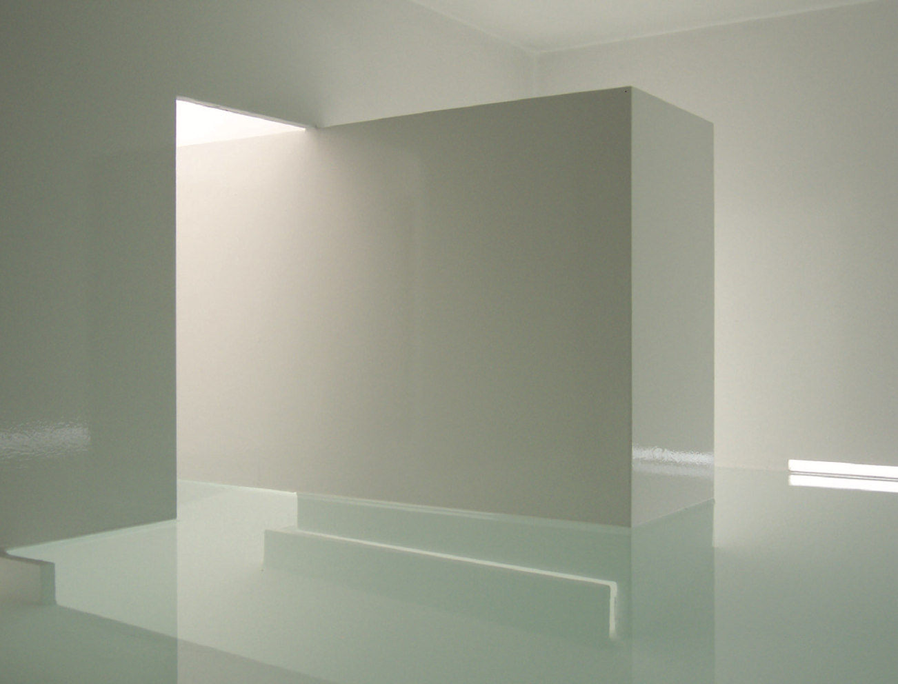 Espacio interior minimalista con paredes y superficies blancas, incluyendo un muro en el centro y luz natural que entra por una abertura en el techo.