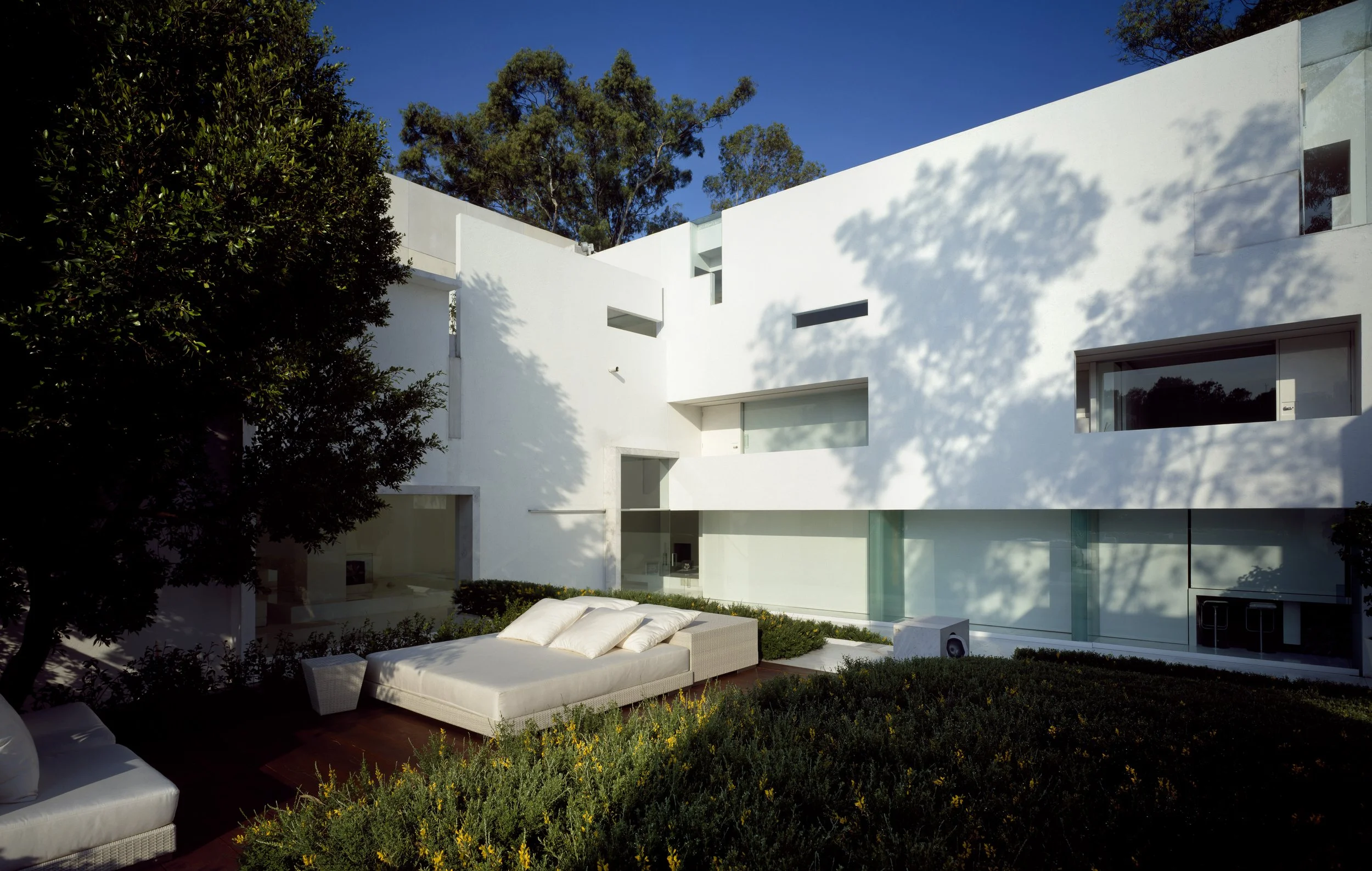 Casa moderna de paredes blancas, con ventanas rectangulares, rodeada de plantas verdes y árboles, con una zona de descanso con sillones blancos y cojines en el jardín, en un día soleado con cielo azul.