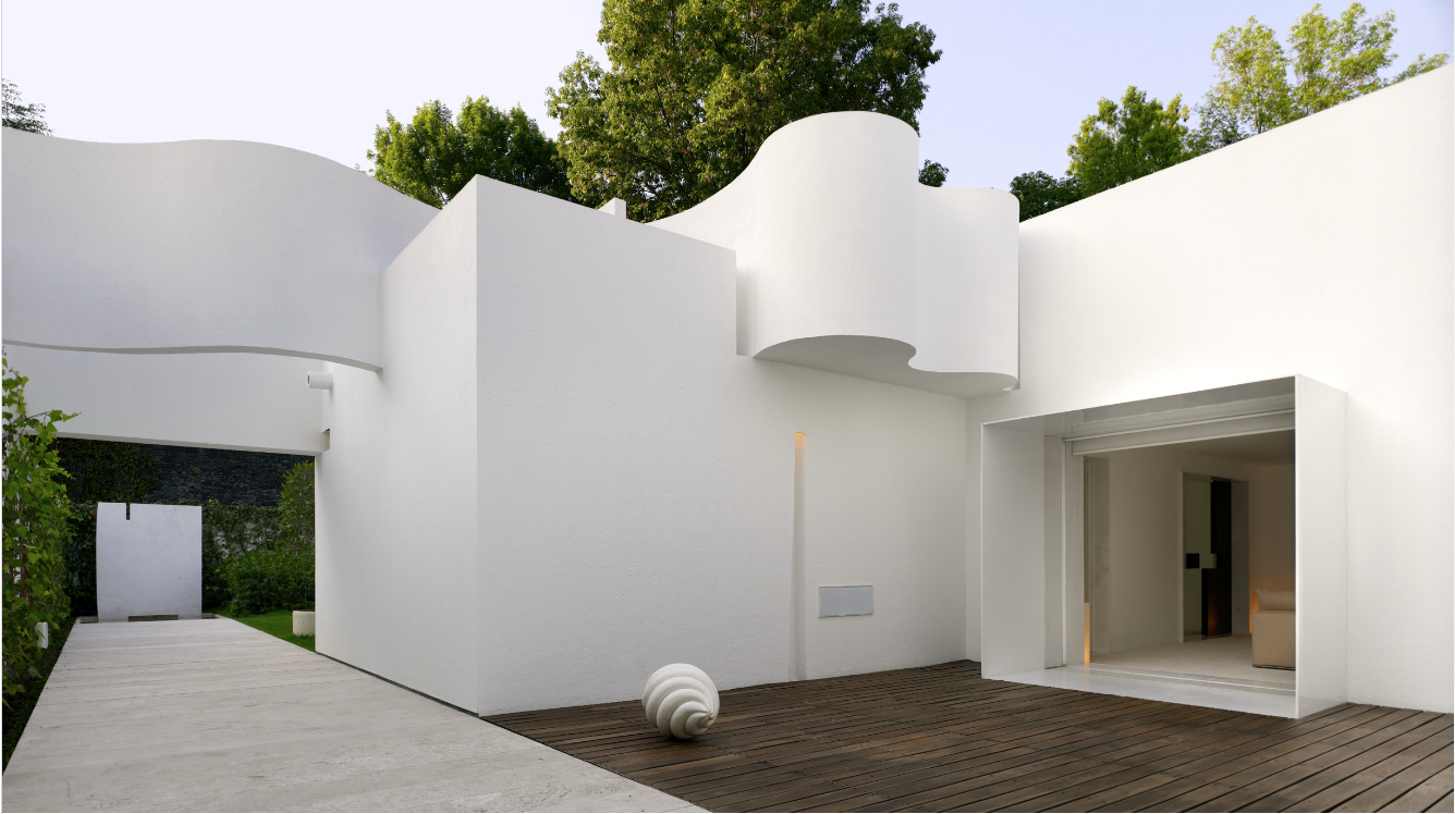 Casa moderna blanca con paredes curvas y un comedor interior visible, rodeada de árboles y con un camino de concreto y madera en el exterior. Eucaliptos 65, Architecture & Design by Rafael Aragonés Caballero & Miguel Ángel Aragonés | Taller Aragonés.