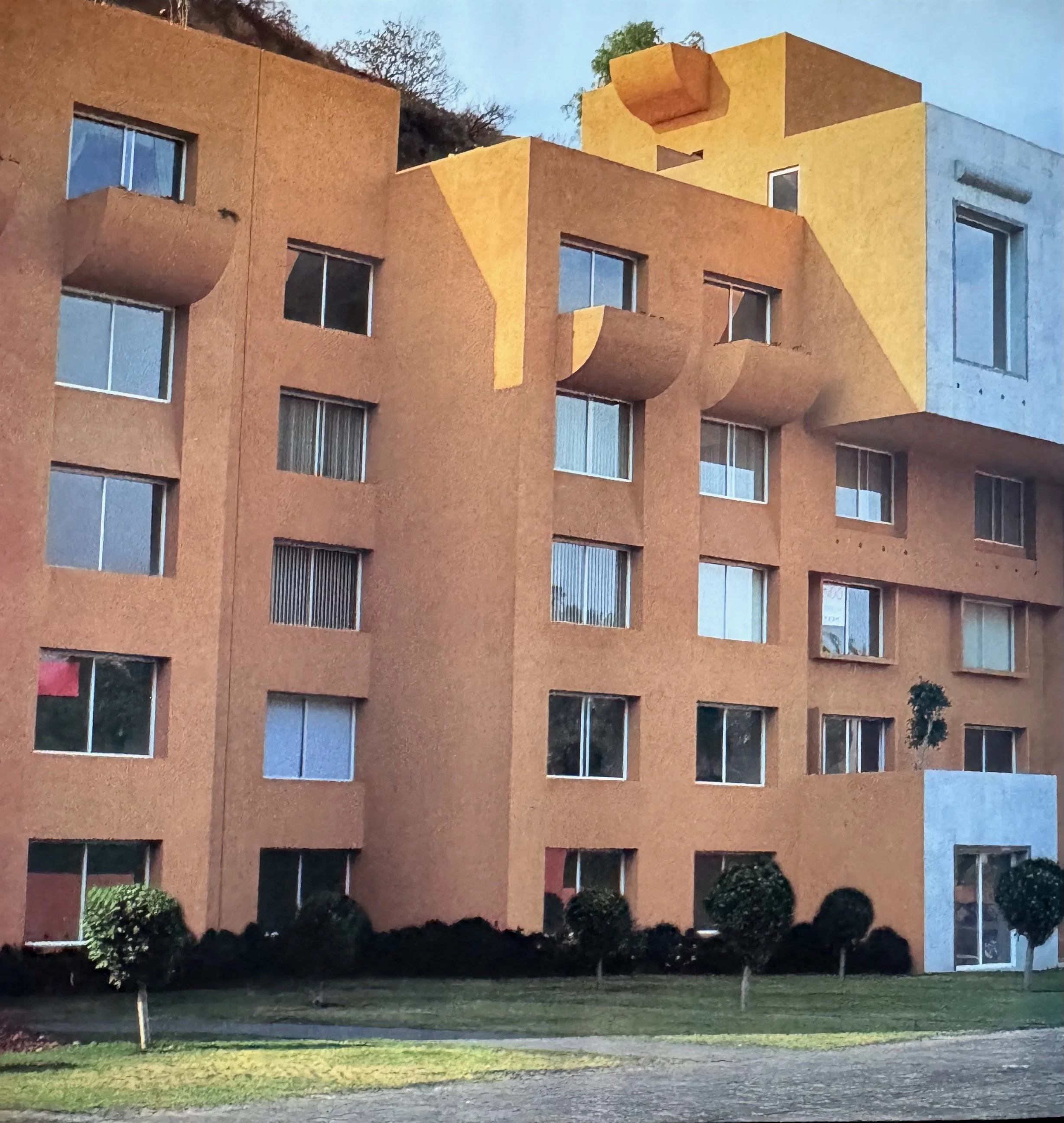 Edificio de apartamentos con fachadas en tonos terracota y amarillo, varias ventanas y balcones, rodeado de jardines y árboles en la tarde.