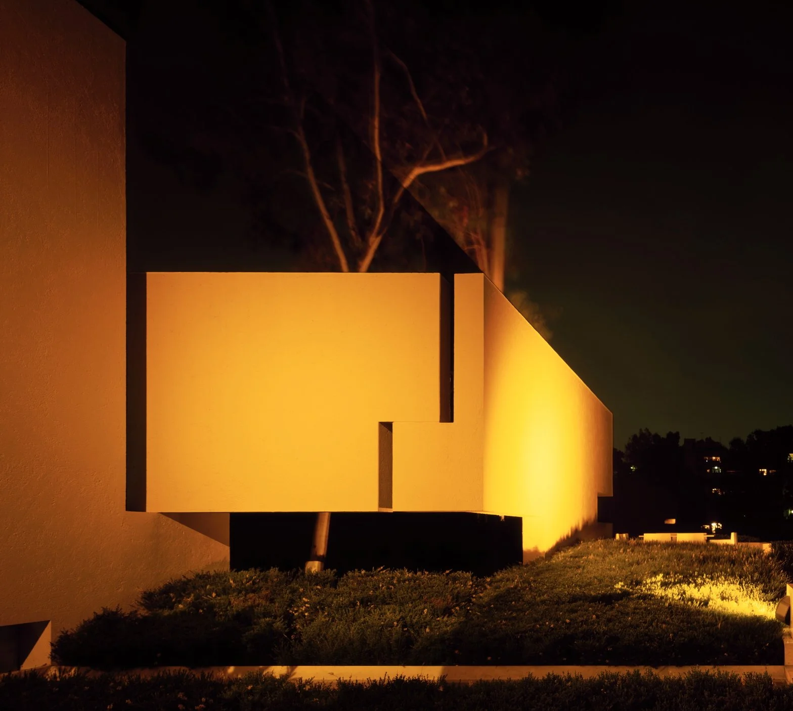 Edificio moderno con paredes blancas iluminadas por luz cálida, sombra de árboles en el fondo, vista nocturna. Architecture & Design by Miguel Ángel Aragonés | Taller Aragonés. Photography by Joe Fletcher