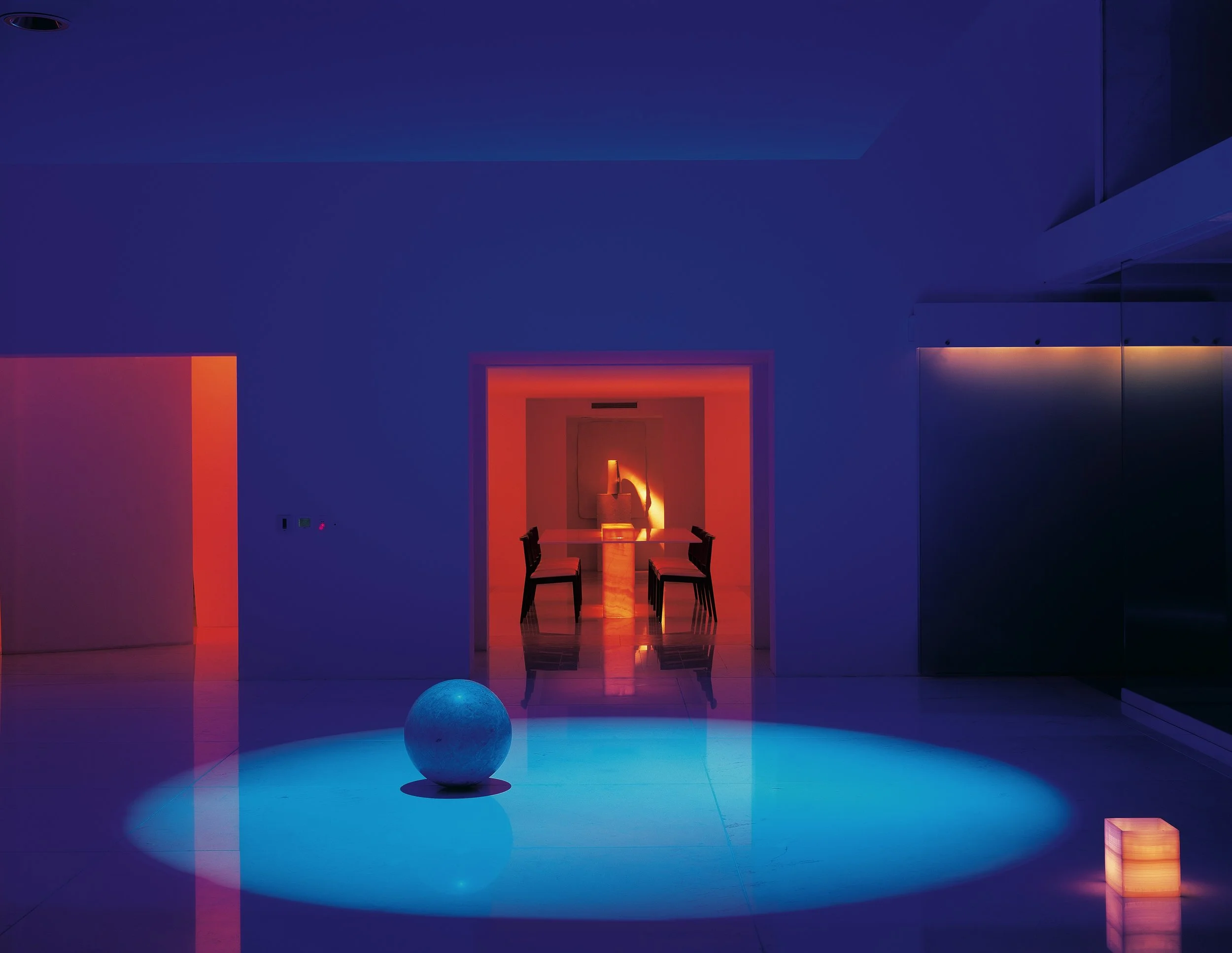 Habitación moderna con luces de colores, mesa y sillas, una esfera en el suelo y paredes con iluminación ambiental. Chromatic architecture in Eucaliptos 65 by Miguel Ángel Aragonés. A study in vibrant tonalities and light, acknowledging the Modernist