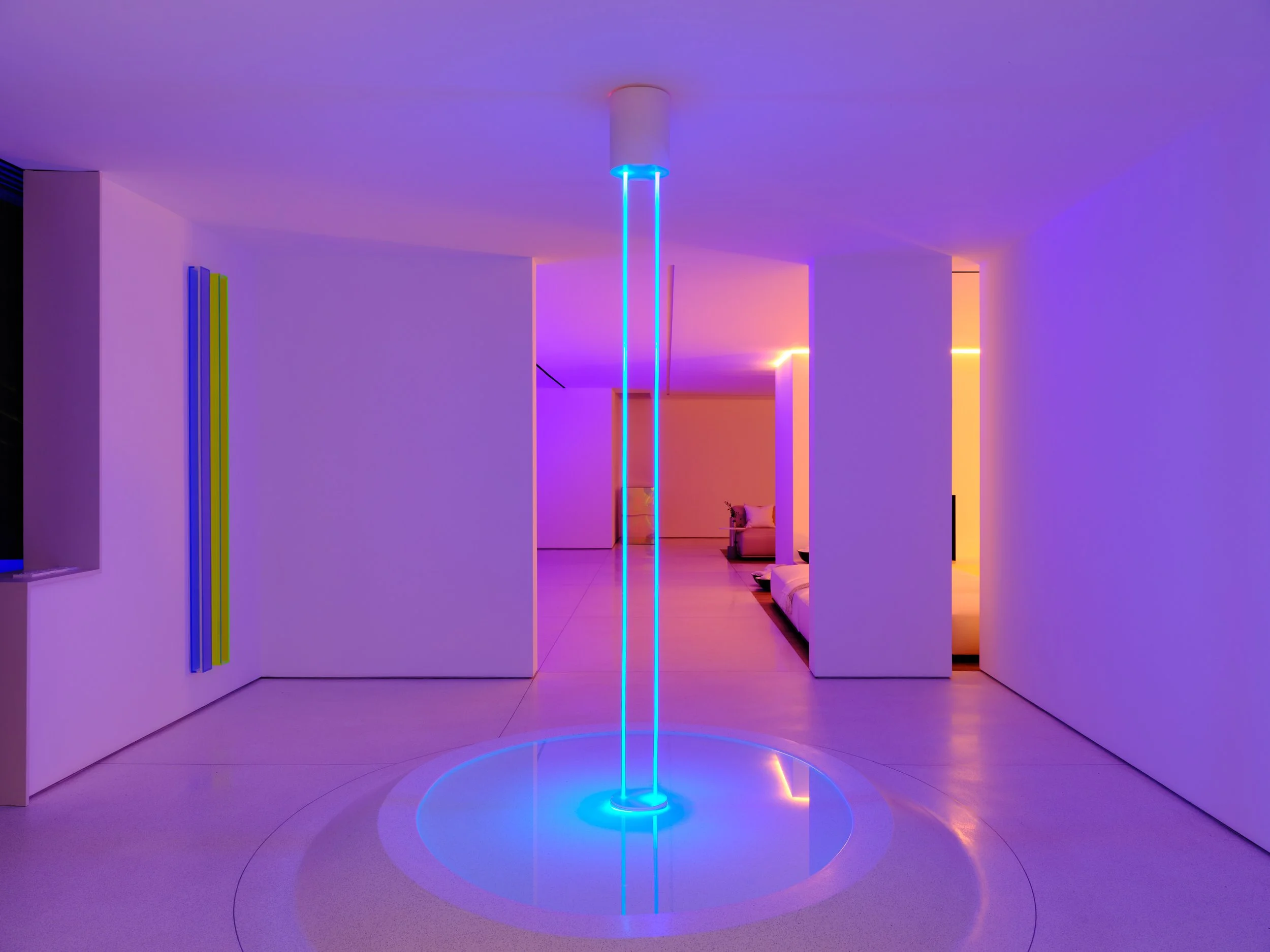 Un espacio interior con iluminación en tonos azules y morados, con una columna de luz azul en el centro y paredes blancas decoradas con líneas de colores llamativos. Architecural Design: Rafael Aragonés, Miguel Angel Aragones. Photo: Joe Fletcher