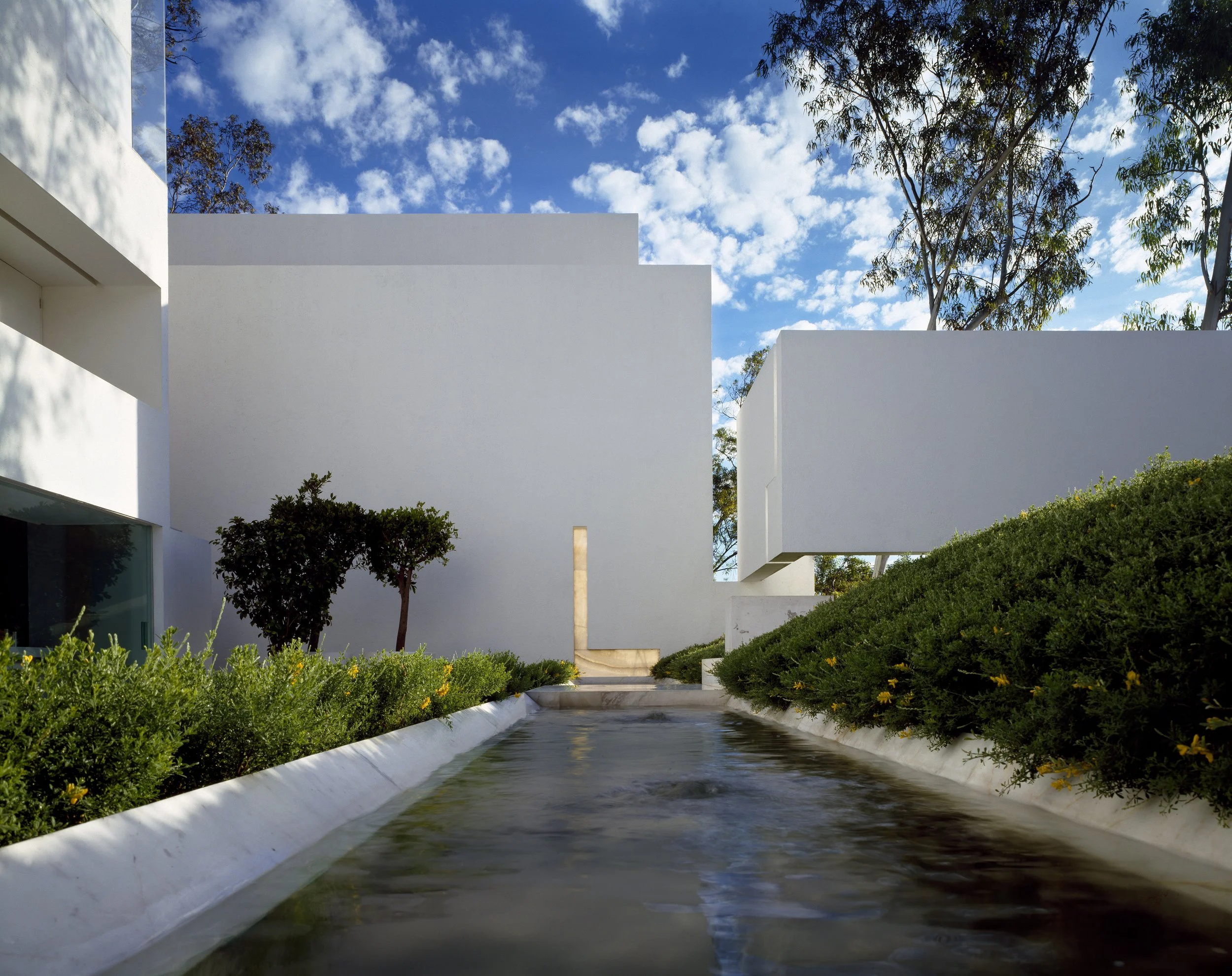 Edificio blanco moderno con formas rectas y ángulos en un cielo azul con nubes, rodeado de árboles y vegetación, con una fuente de agua en primer plano. Architecture & Design by Miguel Ángel Aragonés | Taller Aragonés. Photography by Joe Fletcher