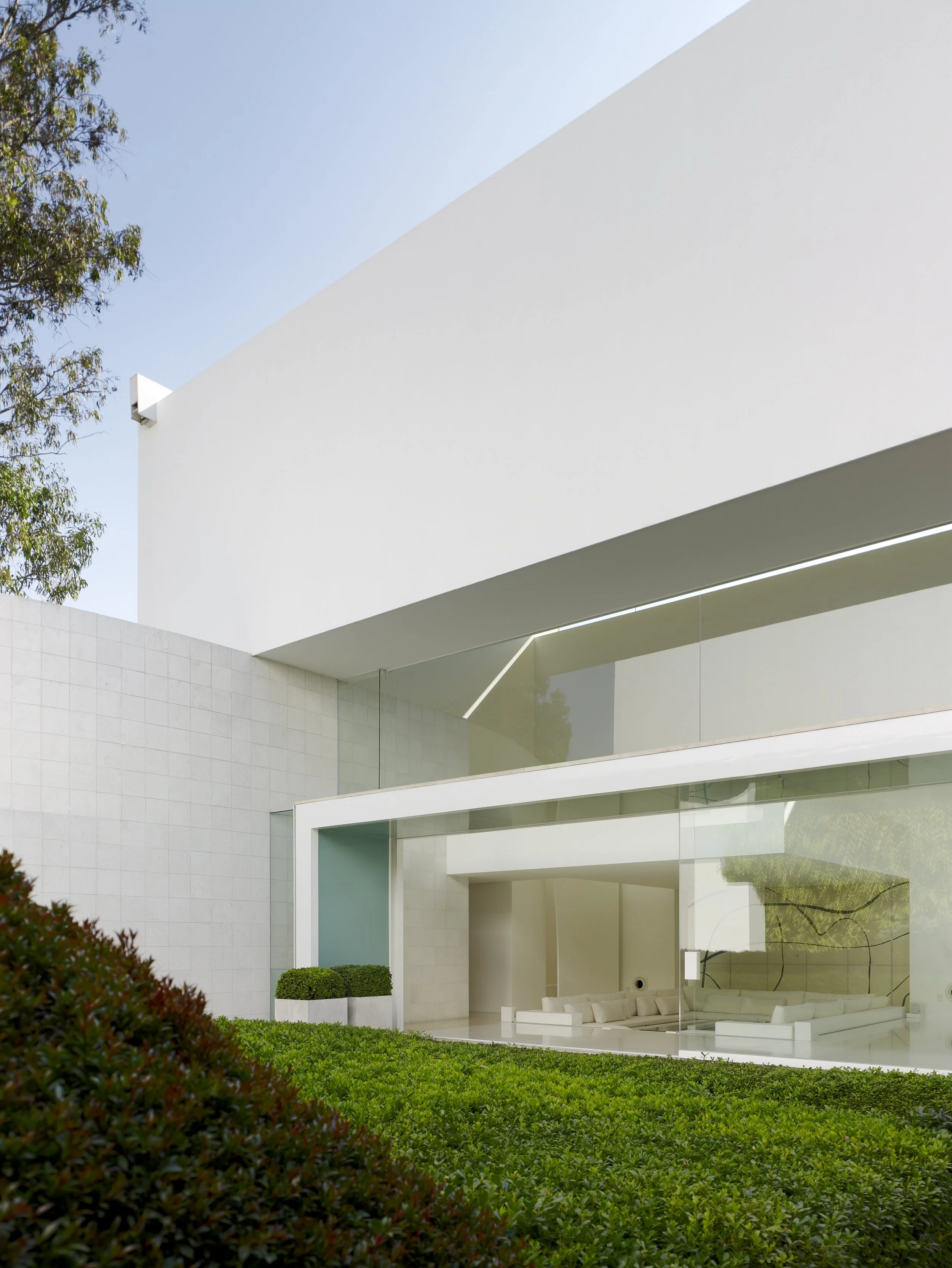 Vista moderna de una estructura blanca con paredes de vidrio y área de jardín con plantas verdes en un exterior soleado. Eucaliptos 65 (2004). Designed by  Miguel Ángel Aragonés. A study in geometric volumes and natural light. Mexican modernism and e