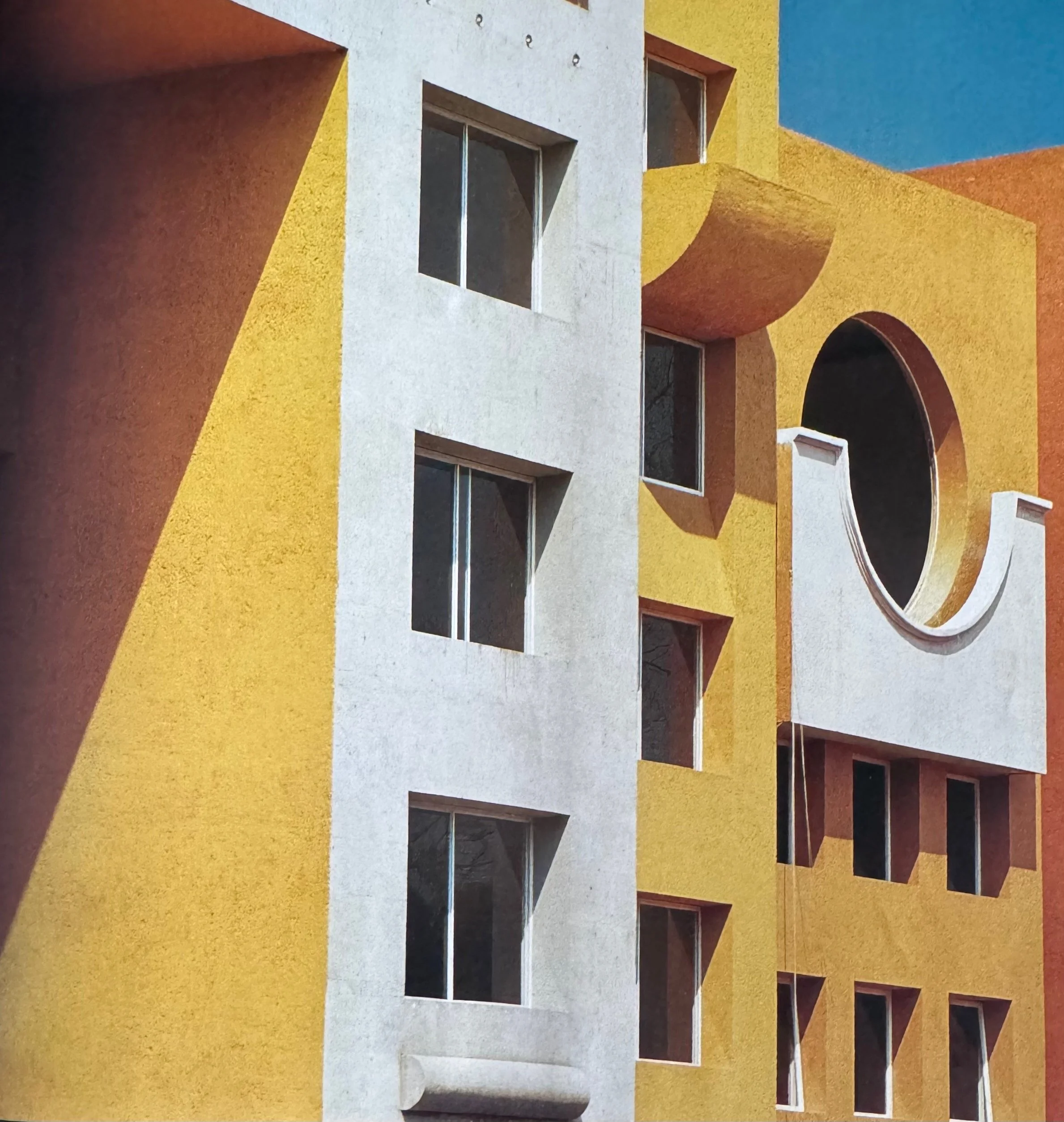 Fachada de un edificio con paredes en colores amarillo, blanco, naranja y azul, con ventanas rectangulares y un arco en la parte inferior derecha.