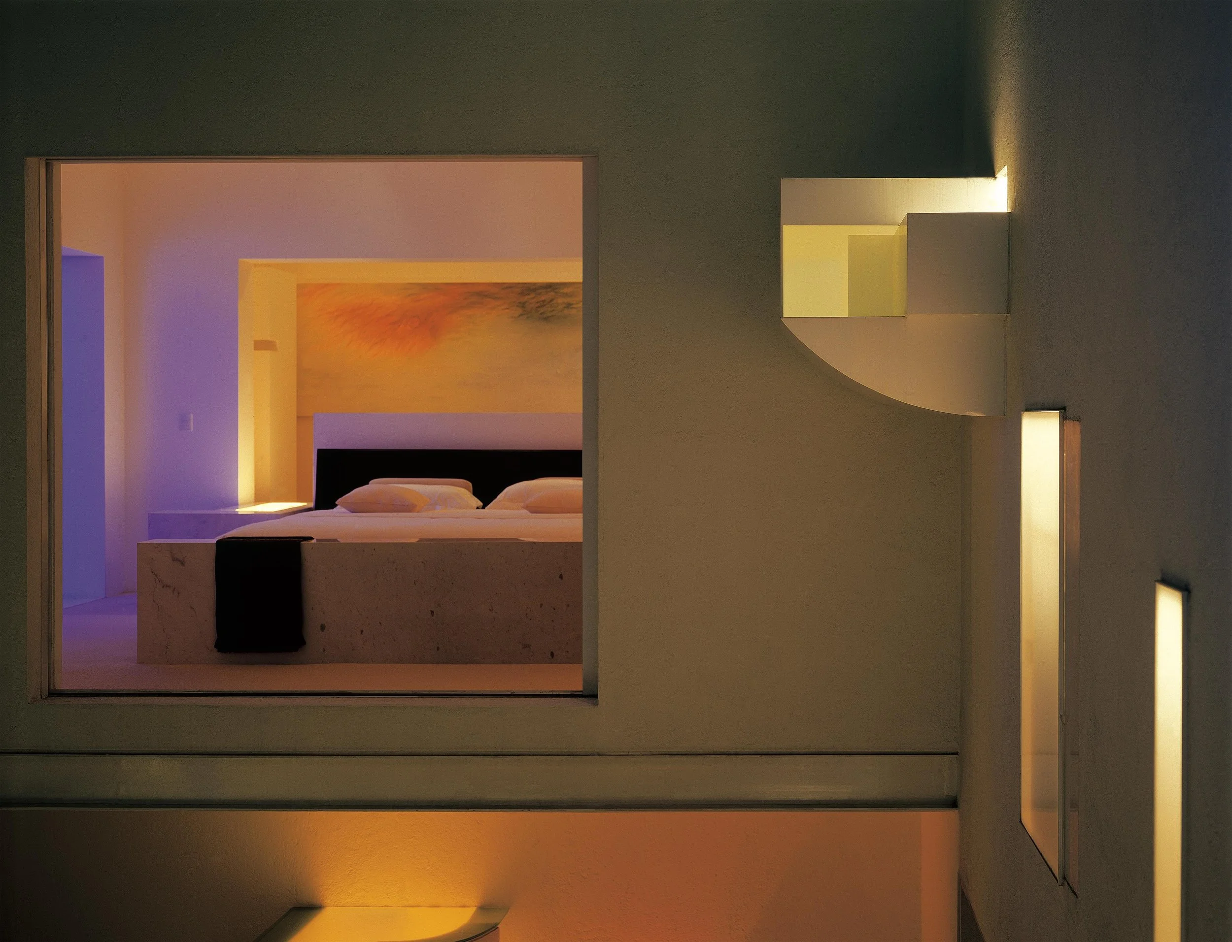 Imagen de una pared con un ventanal que muestra un dormitorio con camas, una pintura abstracta y luz suave, creando un ambiente moderno y minimalista. Eucaliptos 65, Architecture & Design by Rafael Aragonés Caballero & Miguel Ángel Aragonés | Taller 
