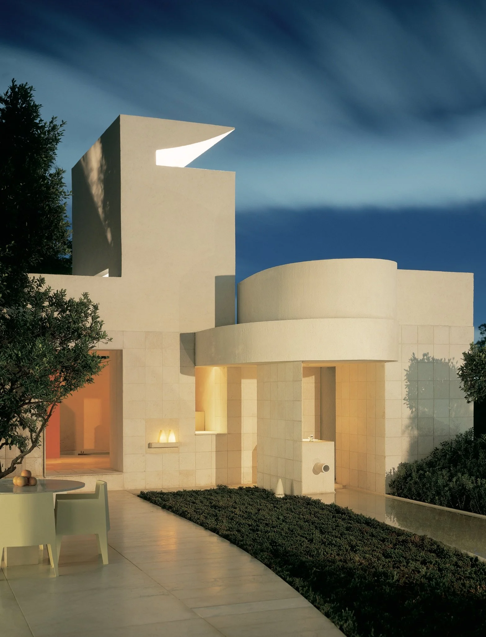 Edificio moderno con formas curvas y rectas, paredes de piedra clara, iluminación cálida, árboles y mobiliario de patio, cielo azul con nubes y árboles verdes.