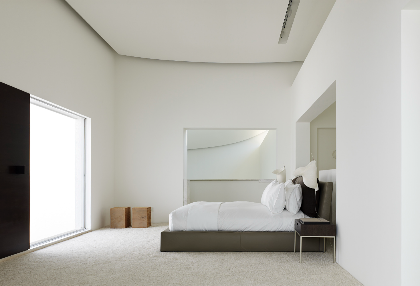 Habitación moderna y minimalista con cama de matrimonio, sábanas blancas y cabecera oscura, con mesa de noche a la derecha, alfombra beige y pared blanca. Eucaliptos 65, Architecture & Design by Rafael Aragonés Caballero & Miguel Ángel Aragonés | Tal