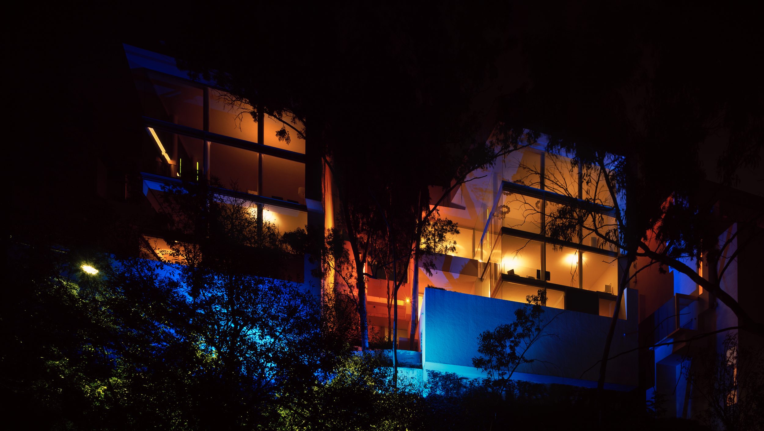 Edificio moderno de varias plantas con ventanas iluminadas en tonos cálidos y fríos, visto de noche con árboles en primer plano.