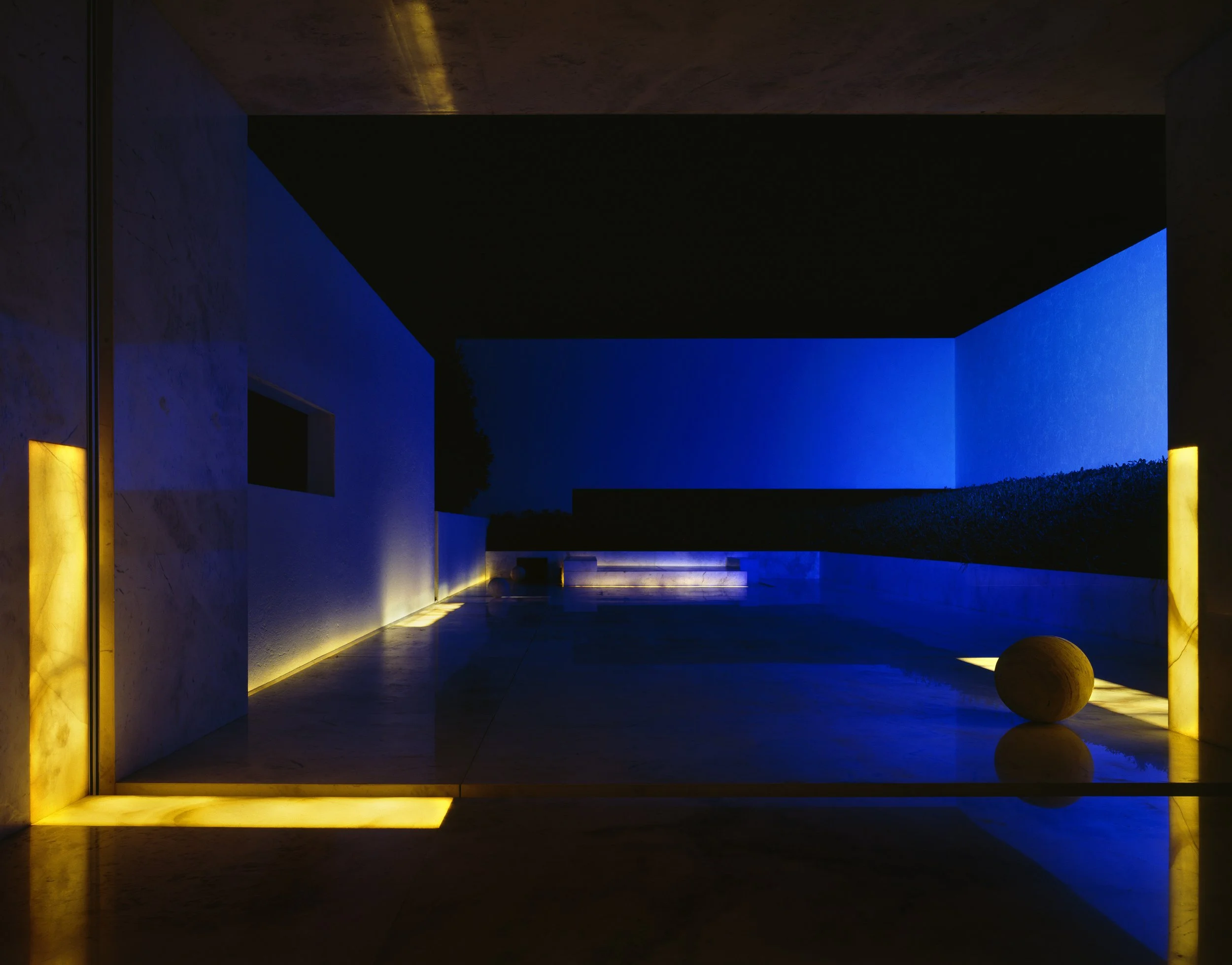 Una estructura moderna con paredes blancas y luces amarillas y azules que iluminan desde abajo y desde arriba, creando un ambiente nocturno y minimalista.
