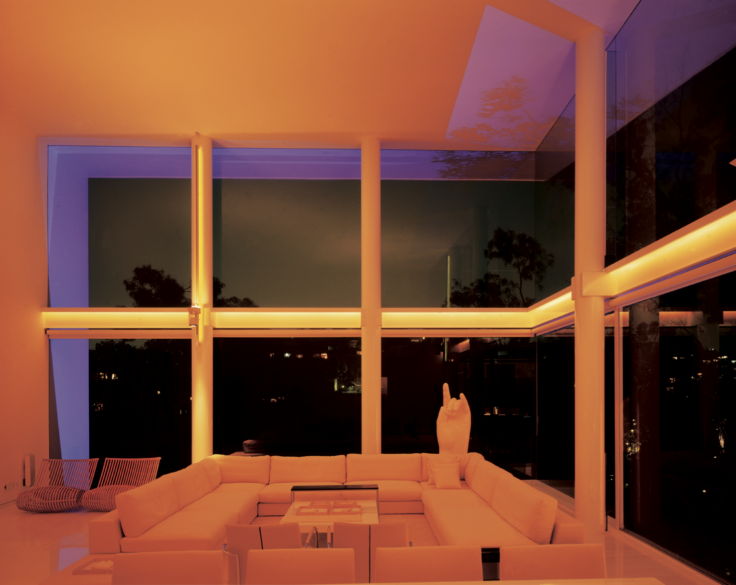 Interior moderno con iluminación de tonos cálidos y grandes ventanas de vidrio que muestran un paisaje nocturno con árboles y luces distantes. Incluye sillones en forma de L y una estatua de mano en el centro.