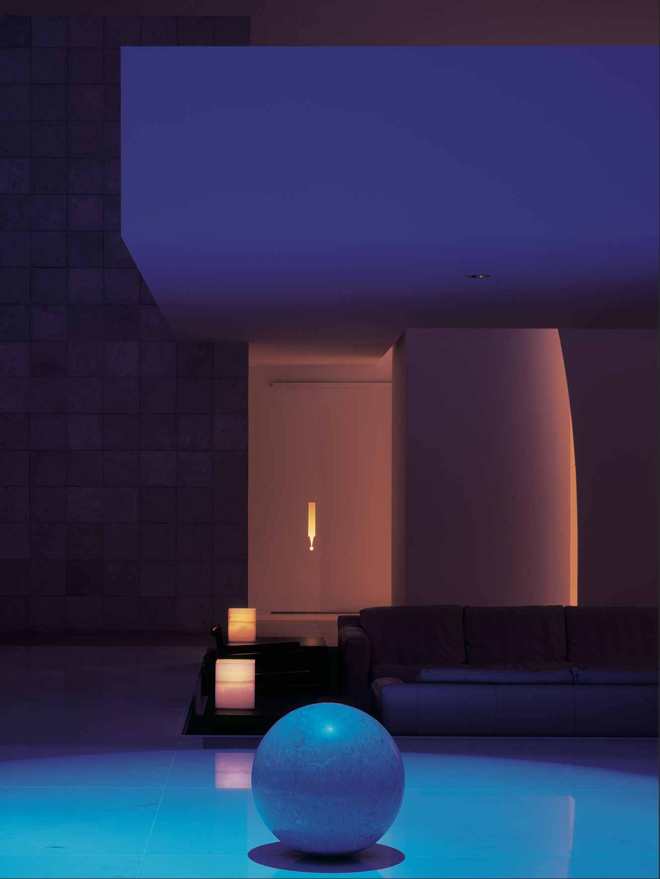 Sala moderna con iluminación tenue, sillón, mesas con lámparas de luz cálida, esfera luminosa azul en el suelo, paredes de colores oscuros y un cielo nocturno en el techo. Chromatic architecture in Eucaliptos 65 by Miguel Ángel Aragonés. A study in v