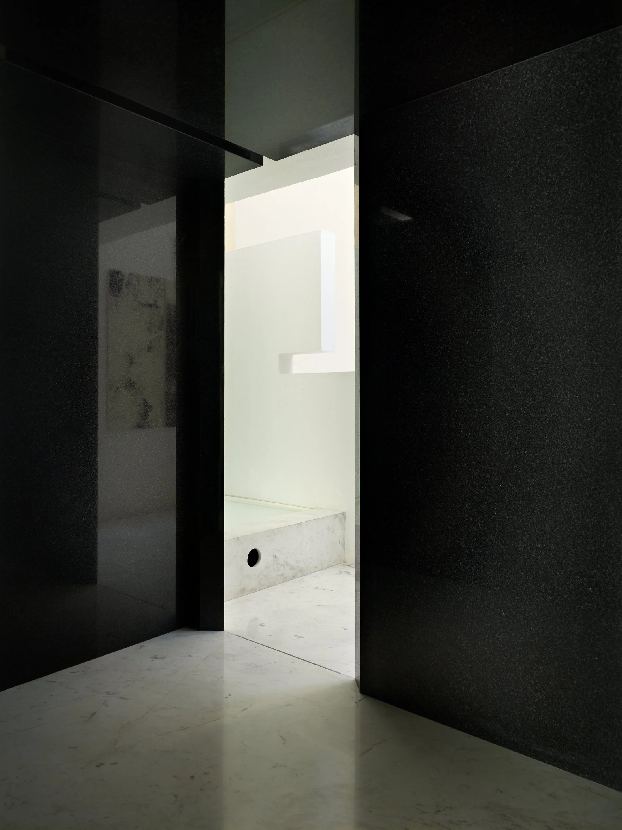 Vista de un pasillo con paredes de color negro y piso de mármol blanco, con una iluminación natural que entra por una abertura en la pared blanca en el fondo.