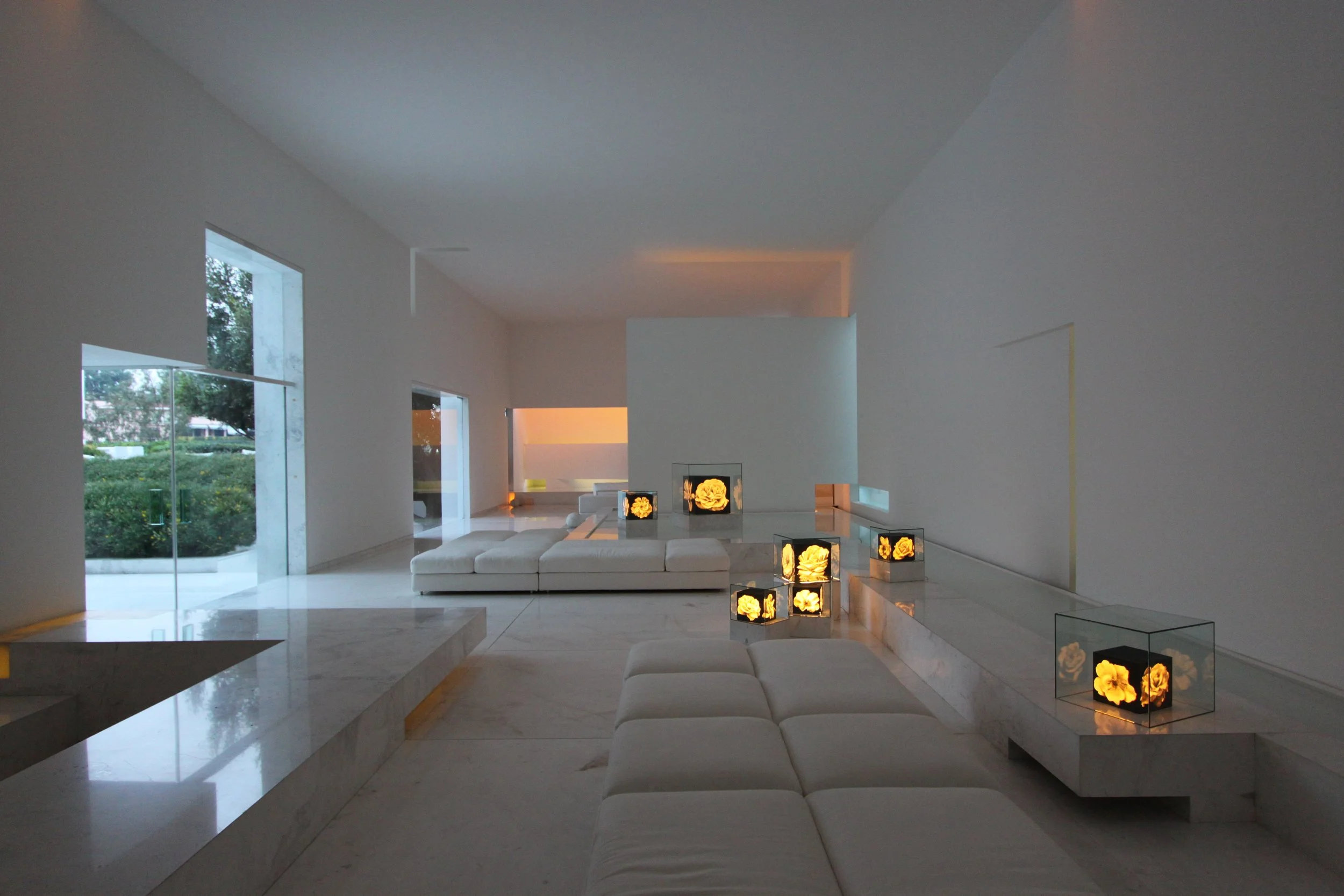 Interior de una sala moderna con paredes blancas, ventanas grandes y muebles blancos, decorada con lámparas en forma de cajas con flores amarillas iluminadas.
