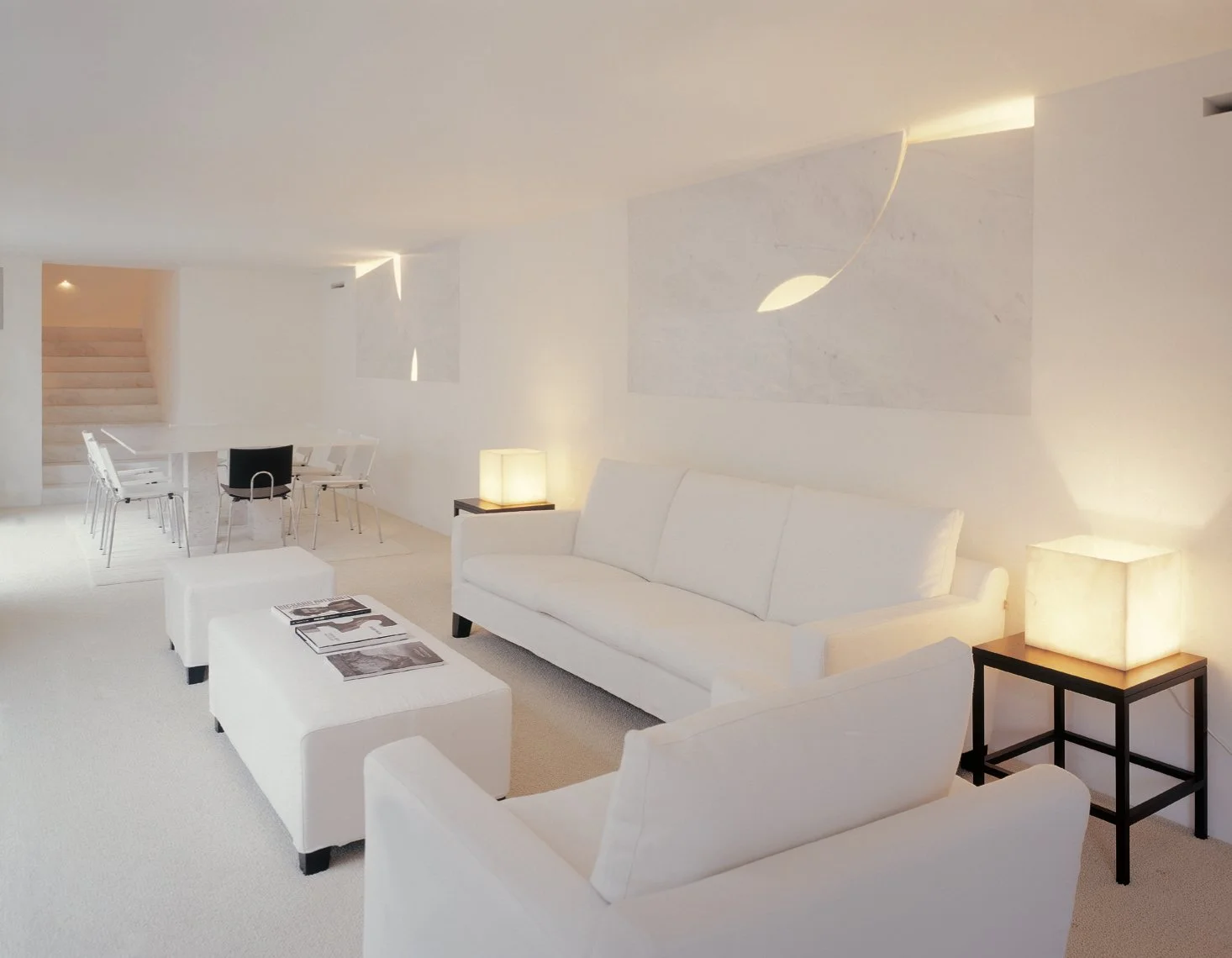 Sala de estar con muebles blancos, lámparas de luz cálida y paredes decoradas con arte minimalista en blanco.