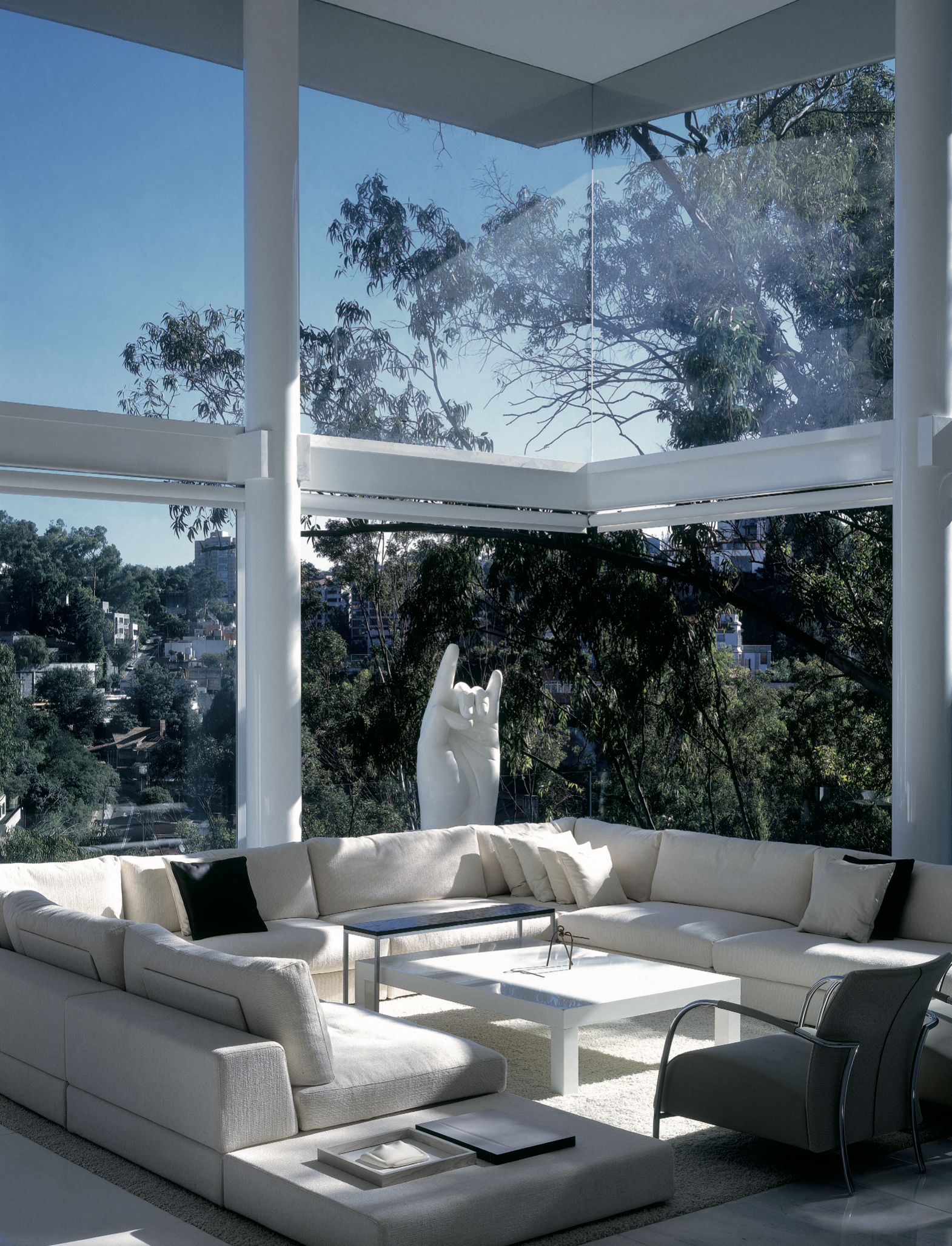 Sala de estar moderna con un gran sofá blanco en forma de L, una mesa de centro blanca, una silla negra y una pared de ventanas de piso a techo que muestra árboles y un cielo despejado.