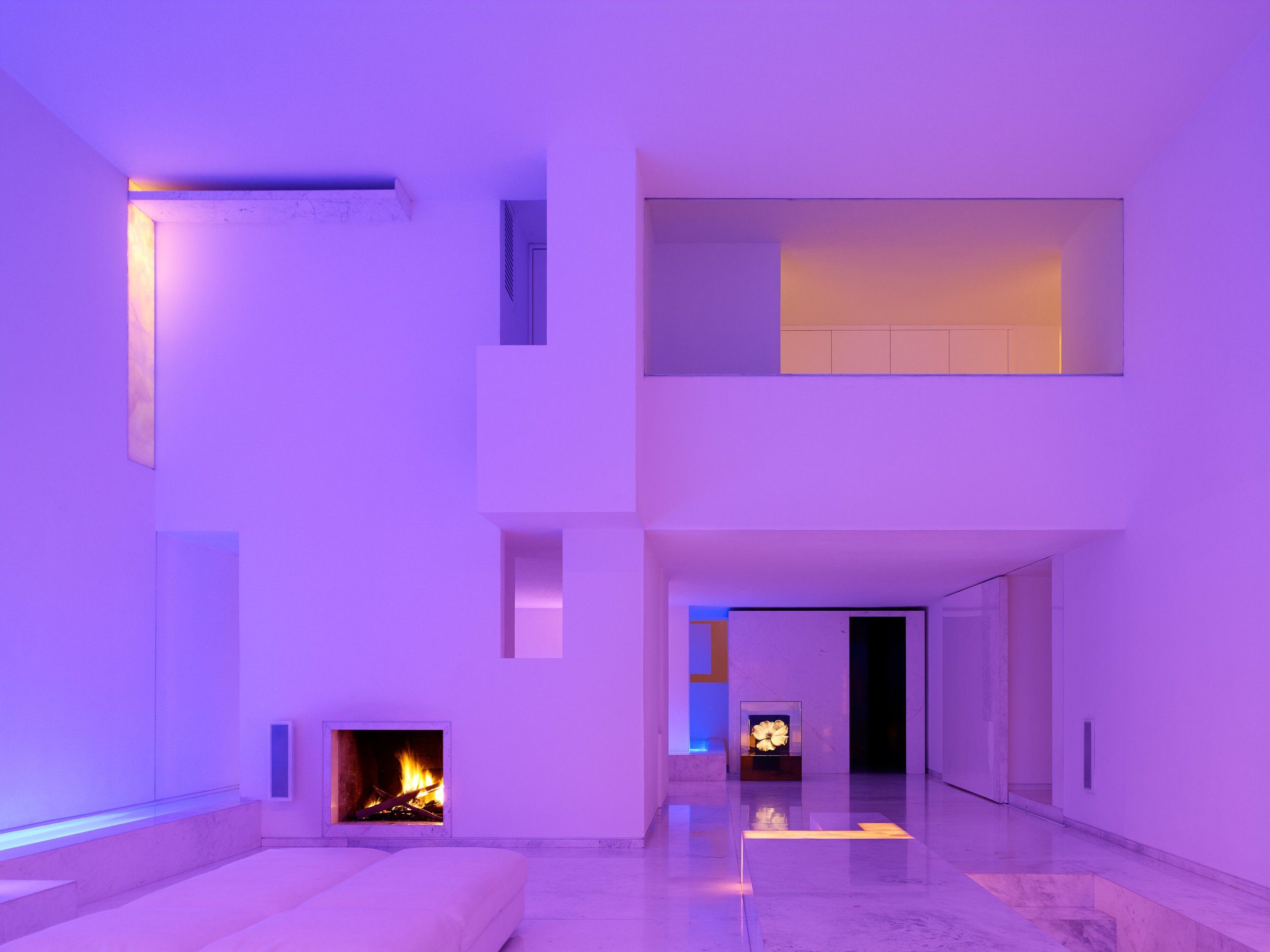 Espacio interior moderno con iluminación de color púrpura y una chimenea encendida en la pared, ventanas grandes y paredes blancas. Architecture & Design by Miguel Ángel Aragonés | Taller Aragonés. Photography by Joe Fletcher